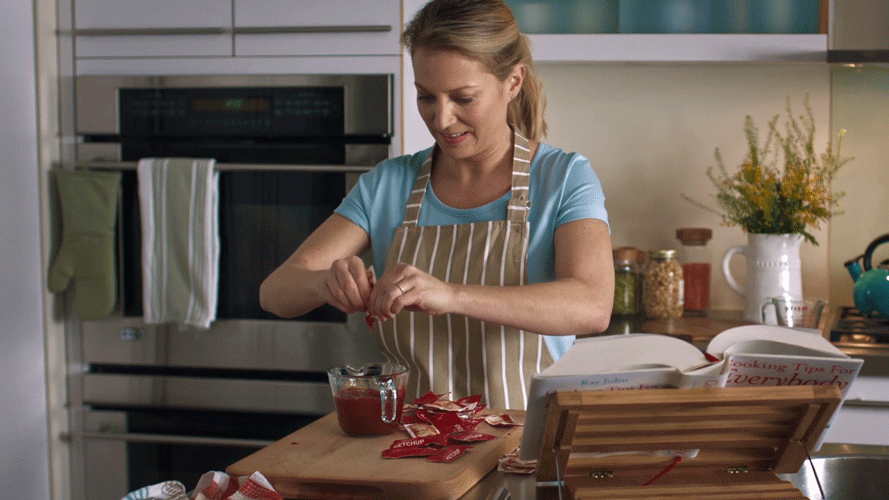A woman peeling red packets of ketchup in a kitchen with various jars and flowers on the countertop.