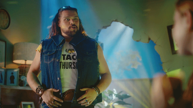 A man with long hair, beard, and mustache wearing a sleeveless denim vest over a T-shirt, standing in a room with a shattered wall, talking to a woman whose face is partially visible.