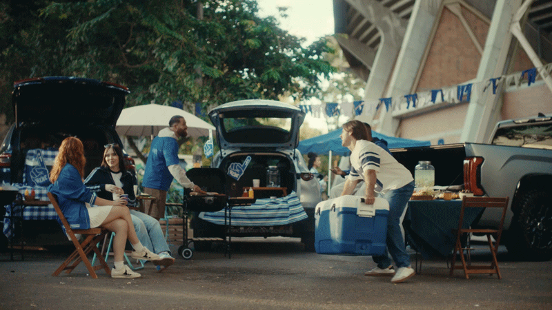 People tailgating at a sports event with food, drinks, and coolers, cars with open trunks behind them.