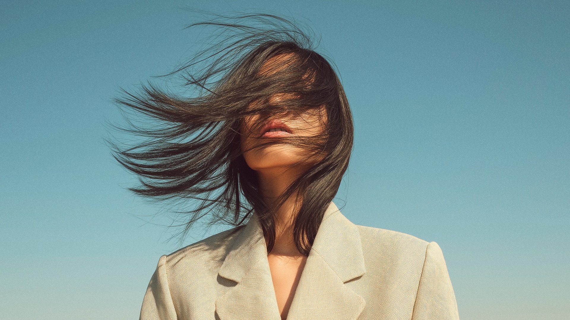 A woman with dark hair blowing across her face, wearing a light-colored blazer, against a clear blue sky.