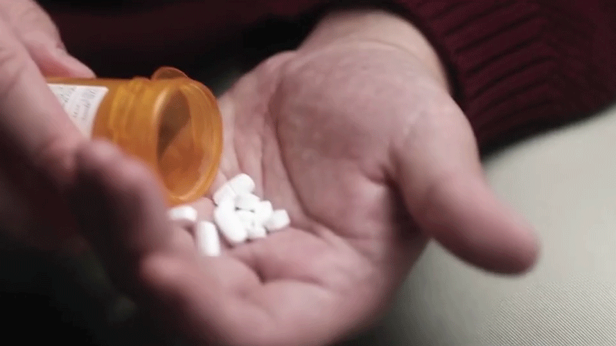 A person's hand holding a pill bottle tilted with white pills spilling into their palm.