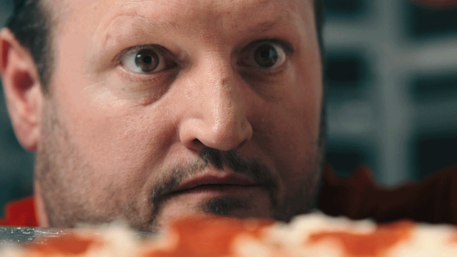 Close-up of a man with a beard and short hair, intently looking at a pepperoni pizza in front of him.