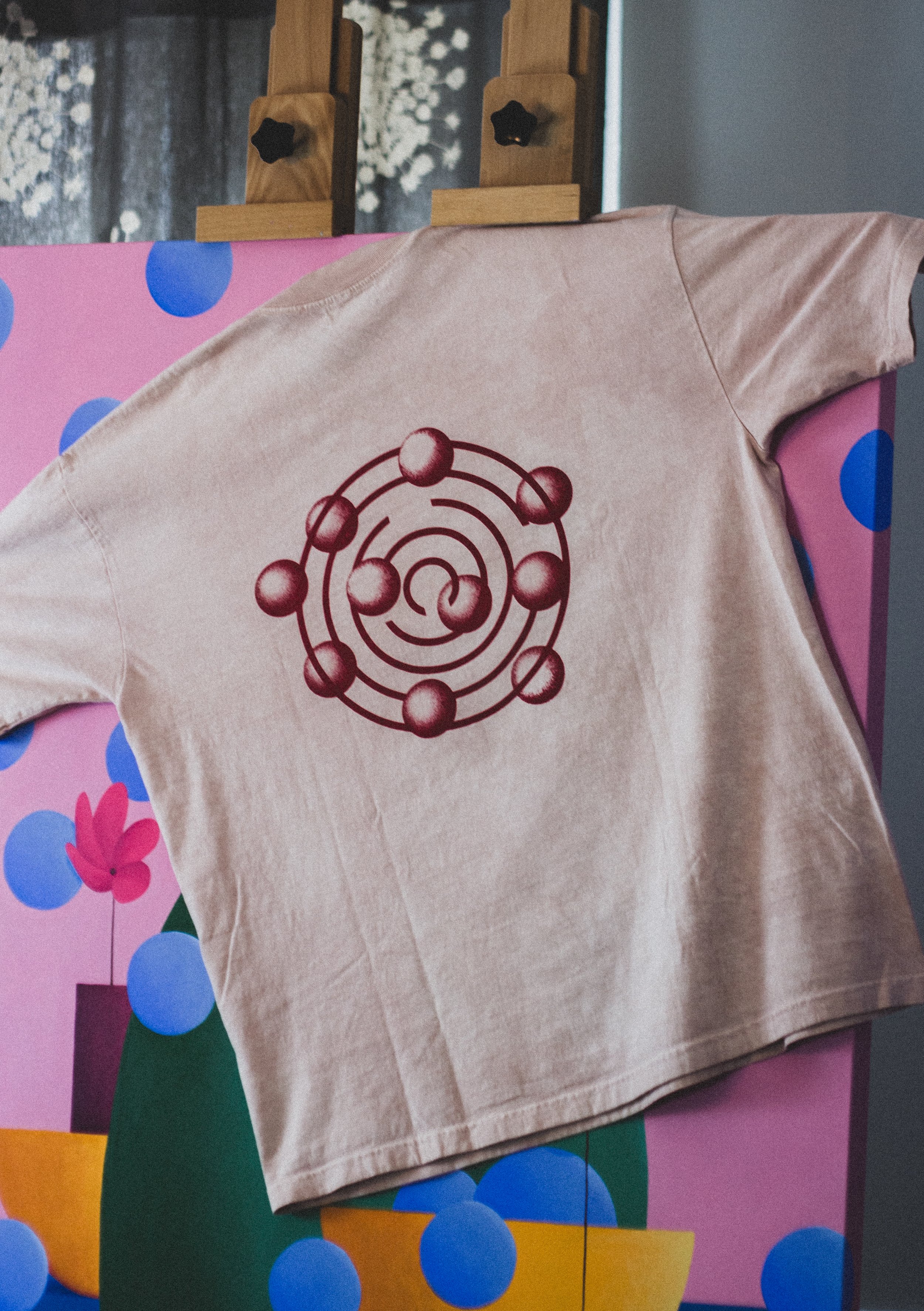 A beige T-shirt with a red atomic structure graphic on the front, draped over a pink and blue polka dot and floral patterned board or surface.