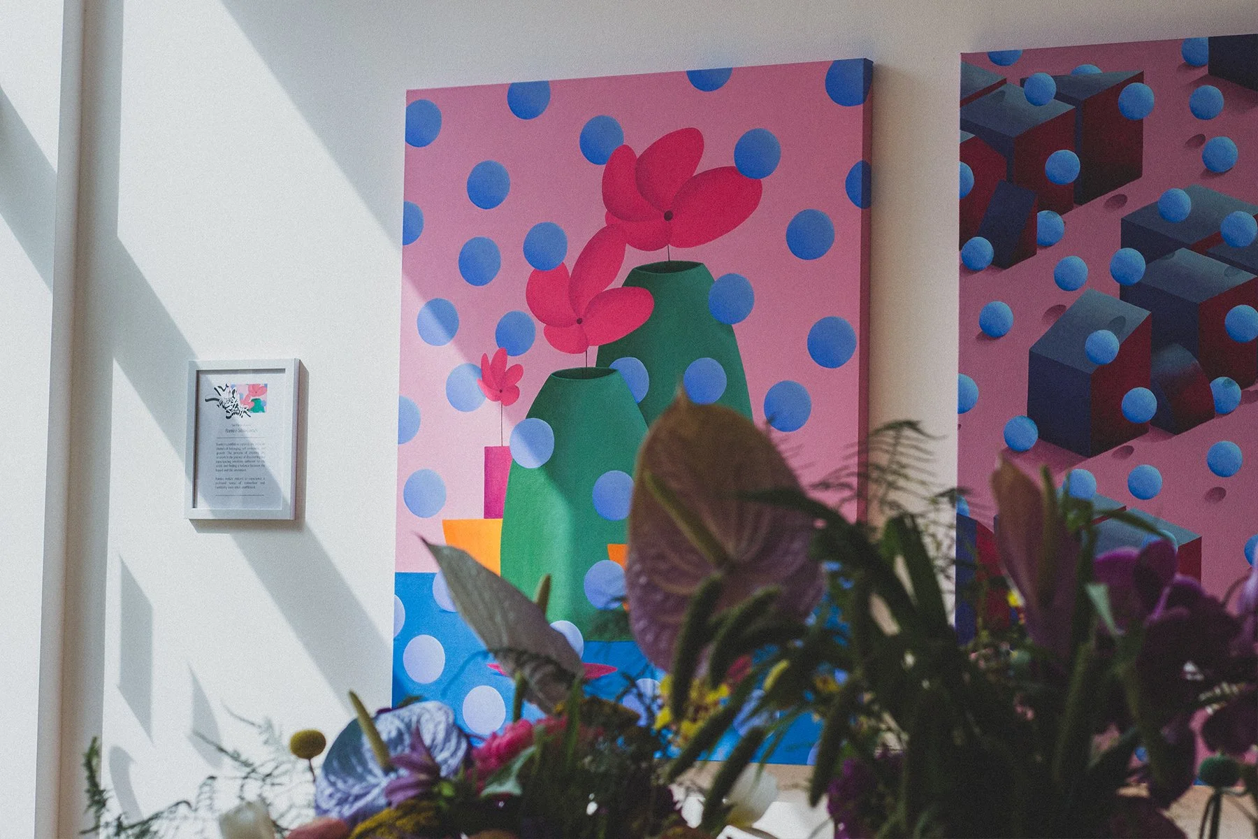 Colorful abstract artwork featuring green vases and pink flowers on a pink background with blue polka dots, displayed in an art gallery with a framed description hanging on the wall.