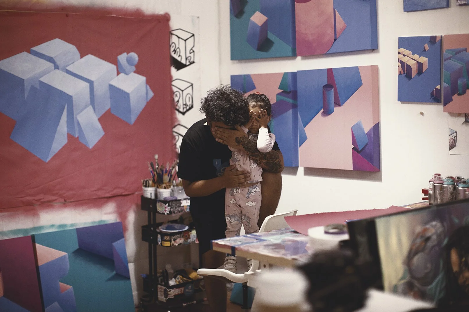 An artist with curly hair and tattoos holding a young girl in pajamas, both covering their faces, in an art studio decorated with colorful geometric paintings and spray paint cans.