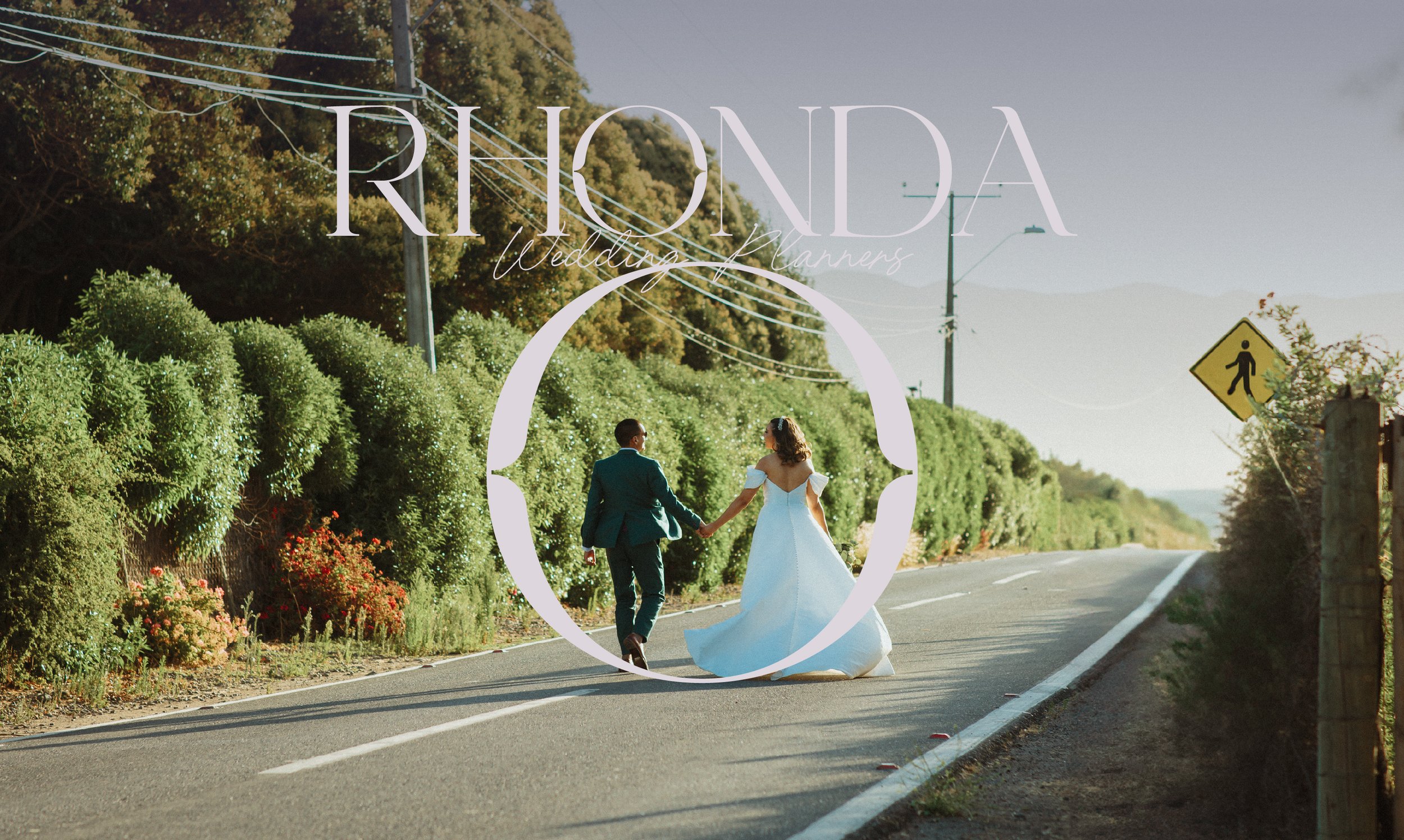 A bride and groom walking hand in hand down a rural road during sunset, with green bushes on each side and a pedestrian crossing sign to the right, overlaid with the text 'Rhonda Wedding Planner.'