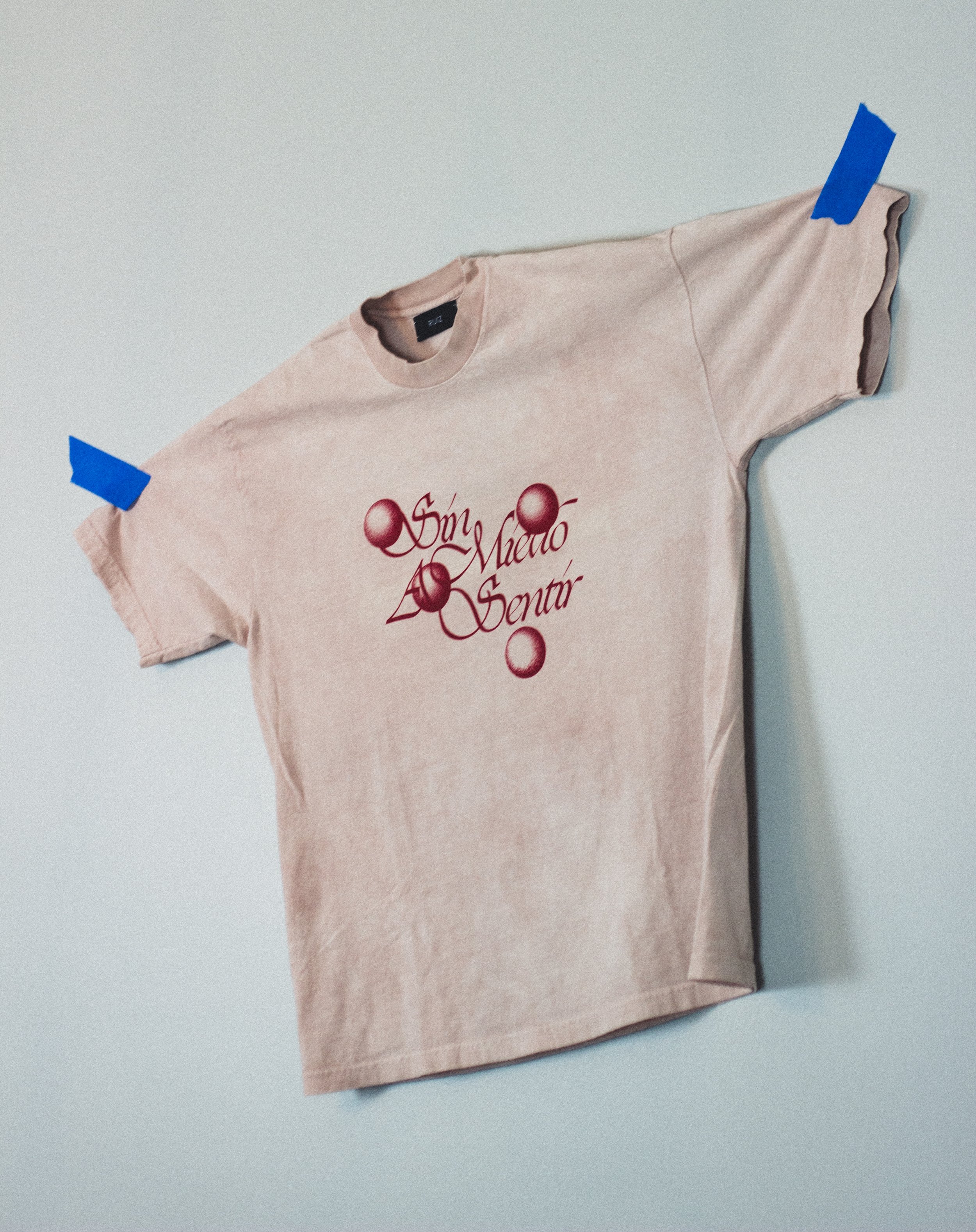 A beige T-shirt with red floral and script lettering design, held up on a plain wall with blue painter's tape on the shoulders.