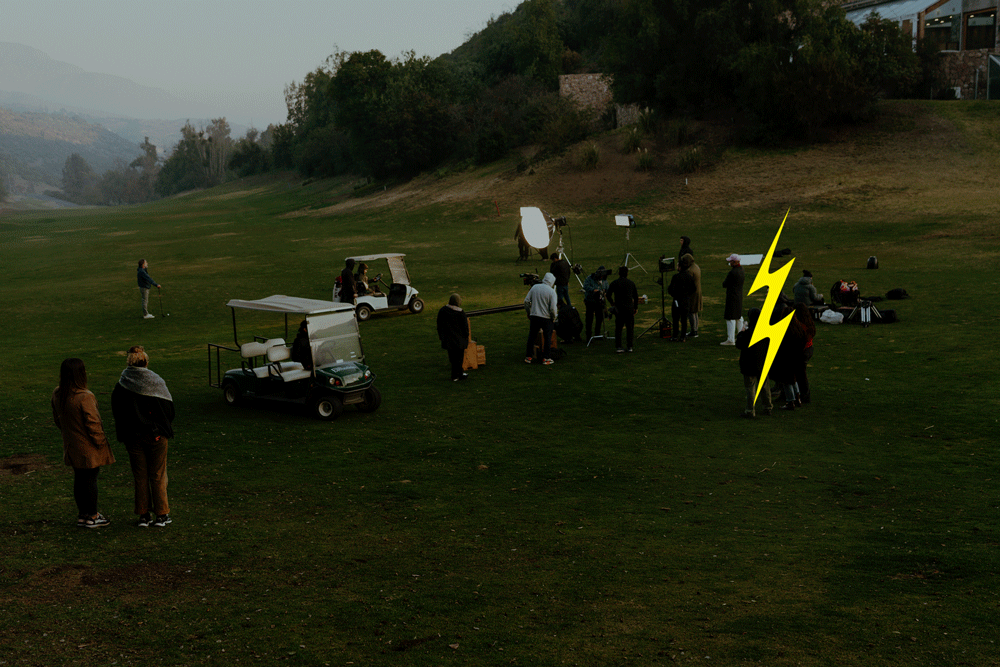 Film crew on a golf course filming a scene with lighting equipment, cameras, and crew members, with two people observing from a distance.
