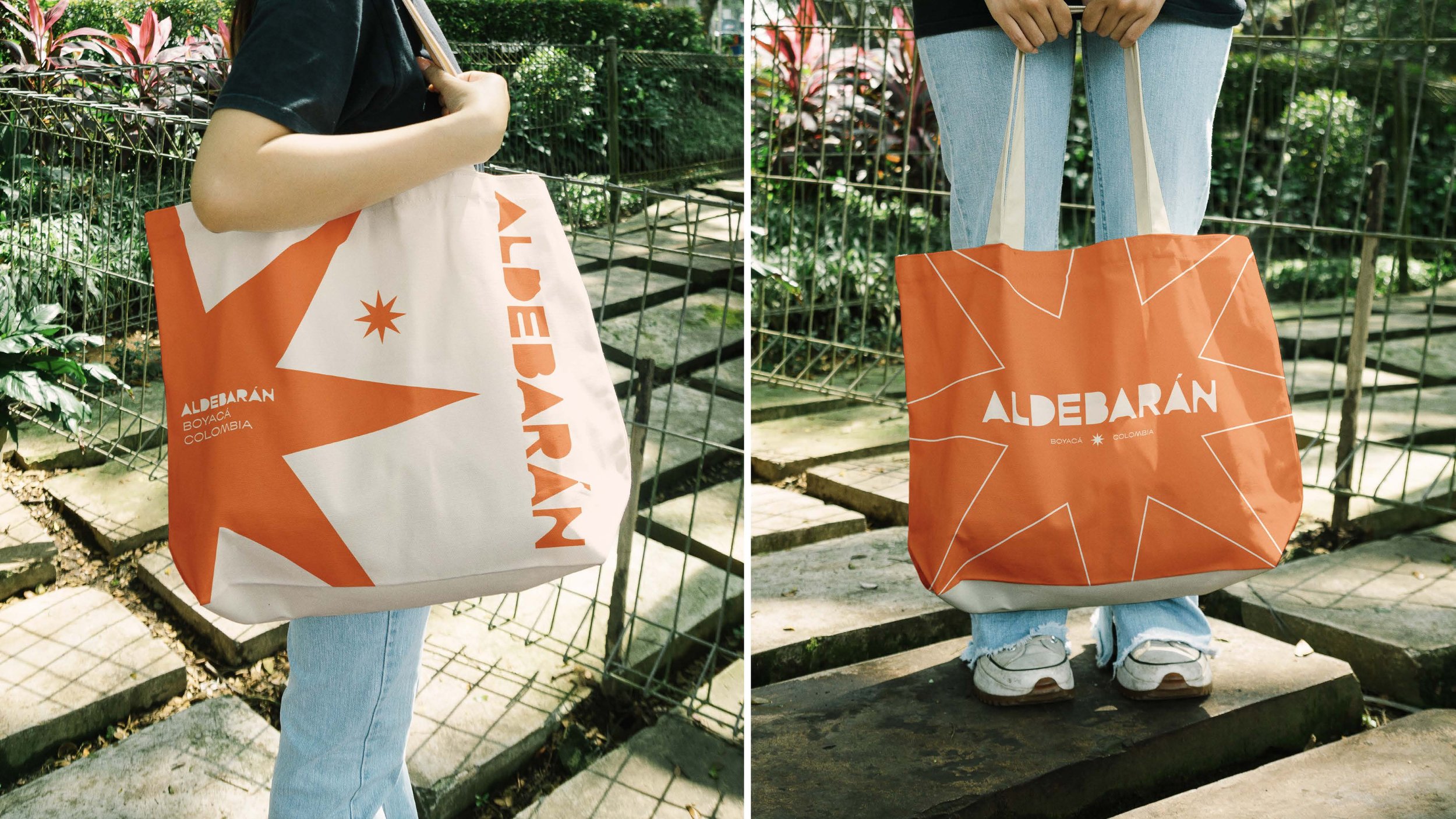Two people holding large tote bags outdoors. The tote bag on the left is white and orange with the text "ALDEBARÁN BOYACÁ COLOMBIA" and a star design. The tote bag on the right is orange with white geometric lines and the text "ALDEBARÁN BOYACÁ COLOMBIA" in white. Both are standing on stone paths near a metal fence with lush greenery in the background.