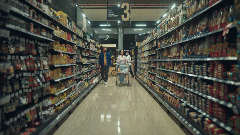 Two people, a man and a woman, walking down an aisle in a grocery store with a shopping cart, surrounded by shelves stocked with various packaged food products.