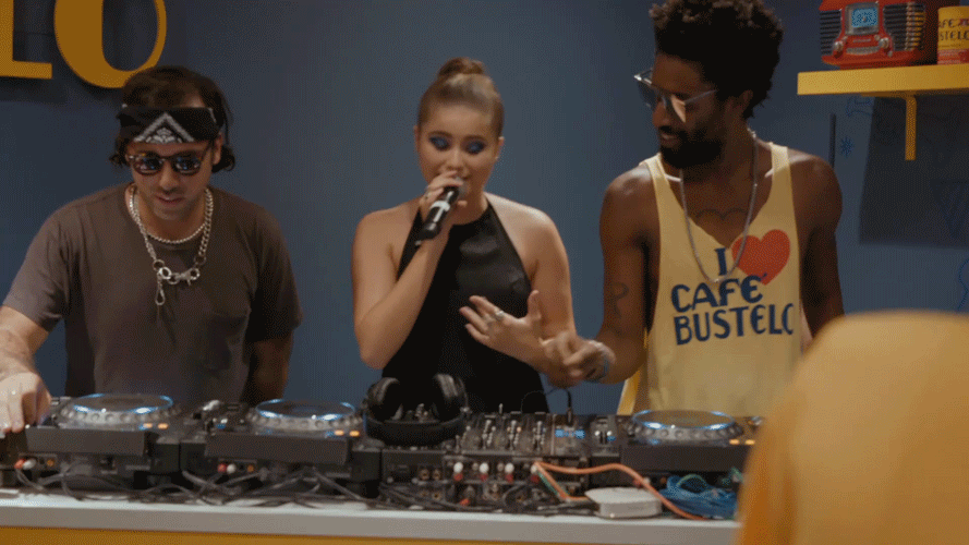 Three people DJs standing behind a DJ setup with turntables and mixer, one is holding a microphone, in a colorful room decorated with objects on yellow and blue shelves.