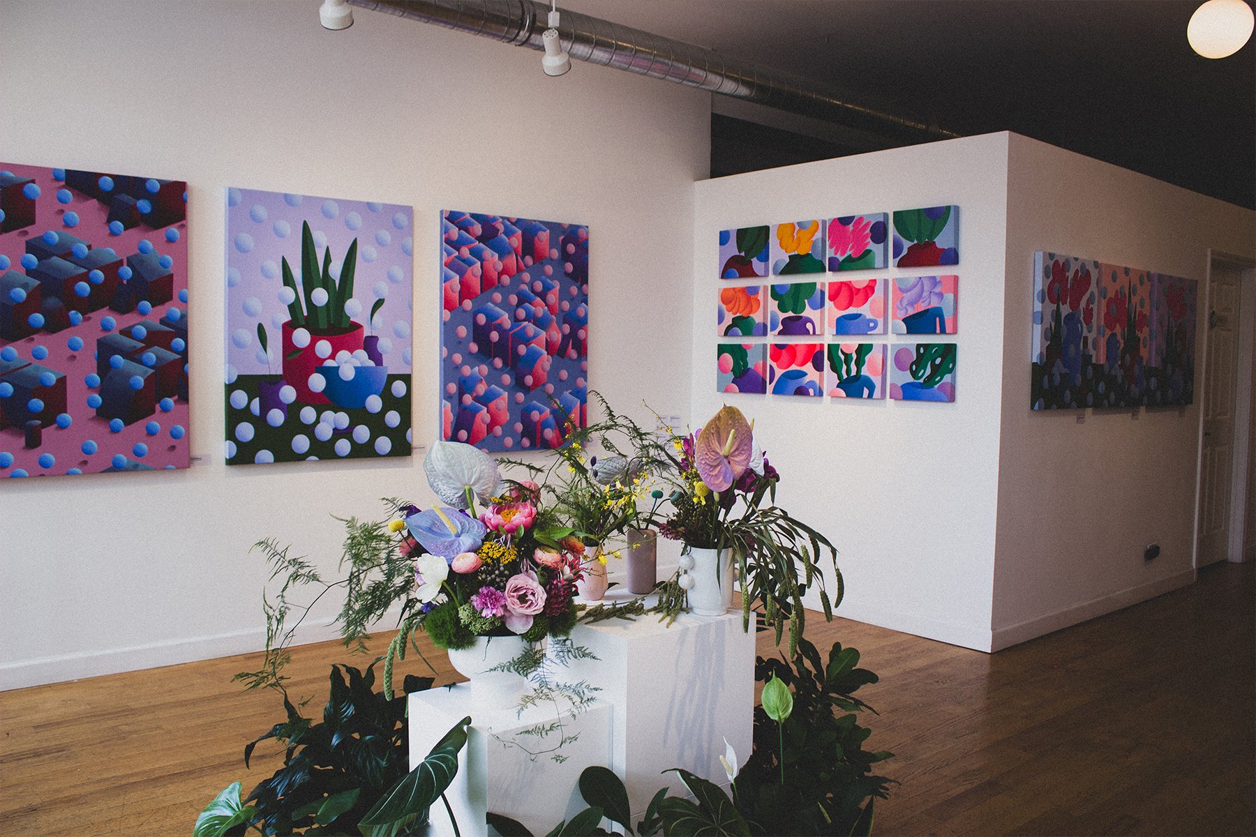 Art gallery with colorful abstract paintings on white walls and flower arrangements on white pedestals in the foreground.