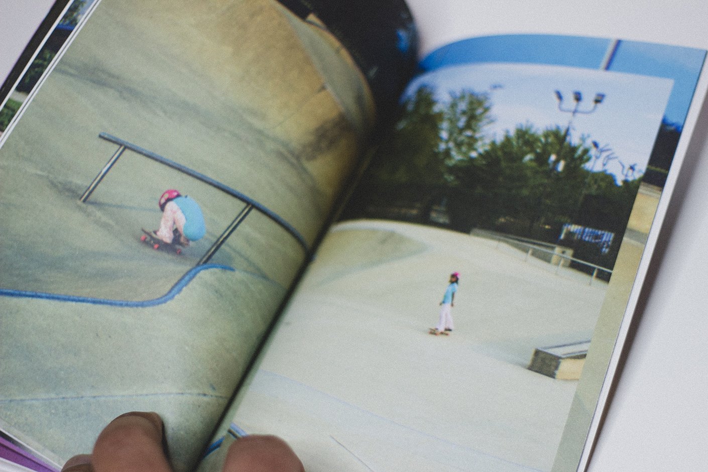 Open photo book showing a skate park with two children skateboarding, one crouching and one standing, both wearing pink helmets.