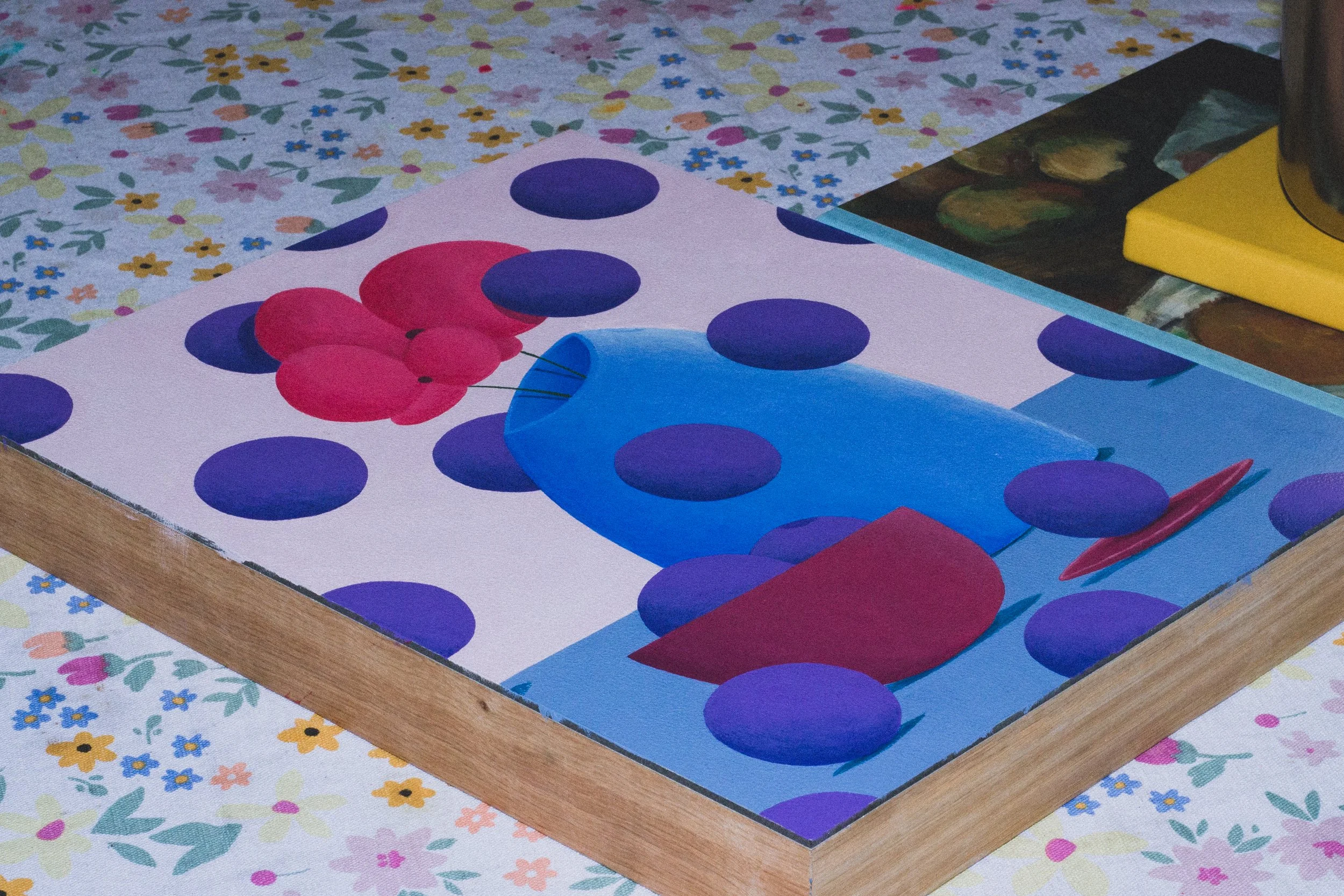 A colorful abstract painting featuring purple dots, red balloons, and a blue fish with a red tail, placed on a wooden frame on a floral tablecloth.