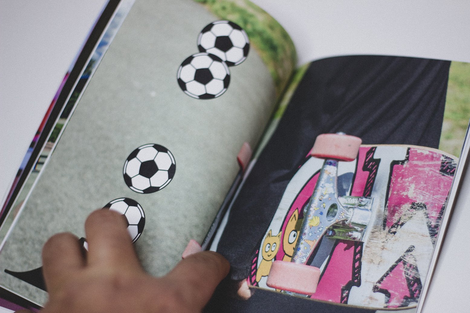A person flipping through a magazine showing a colorful skateboard with pink wheels and a graphic design, and illustrated soccer balls on a grey background.