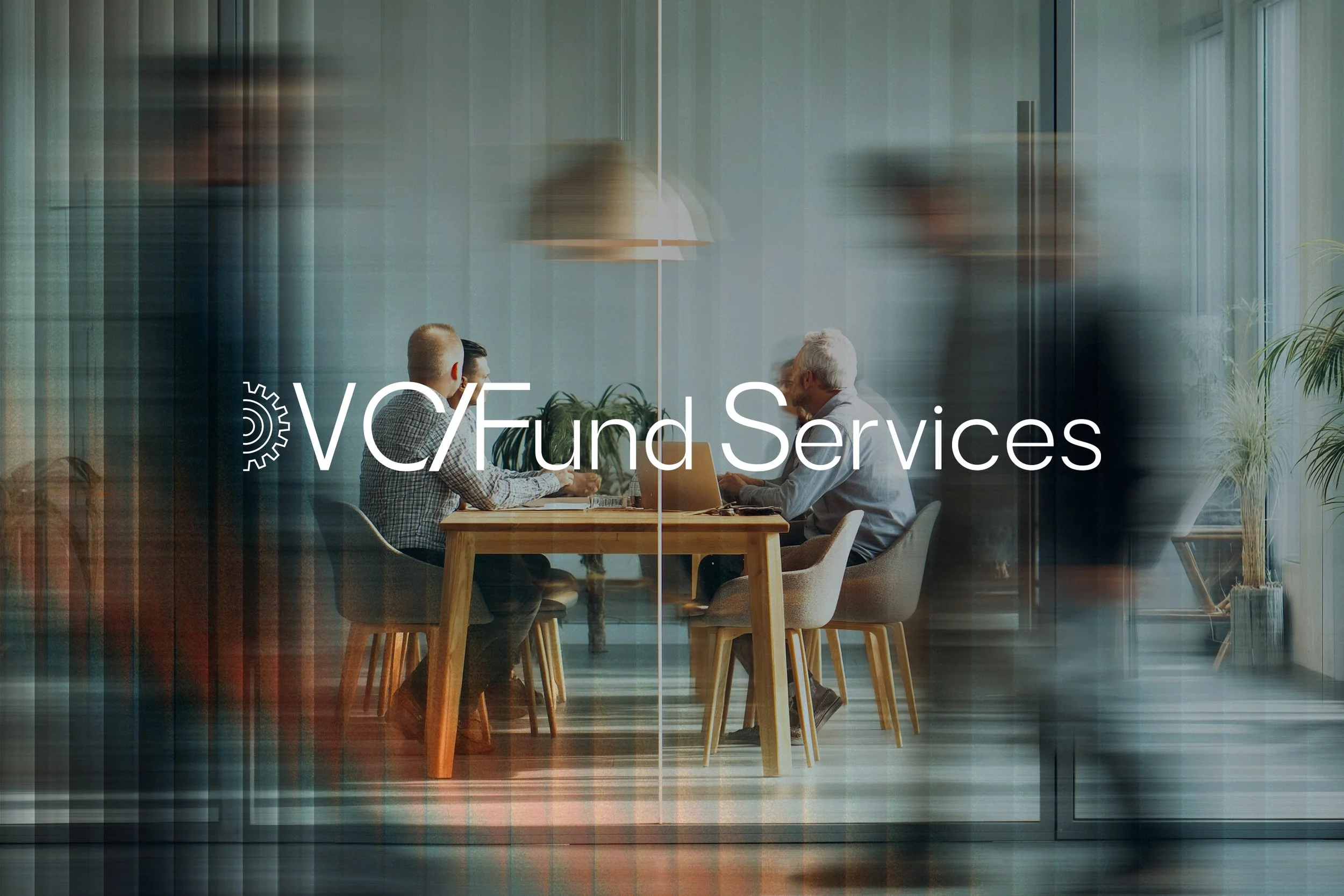 Three people sit at a wooden table in a modern office, engaged in conversation. The scene is viewed through a glass wall with a blurred, reflective effect. Text overlay reads 'VC/Fund Services'.