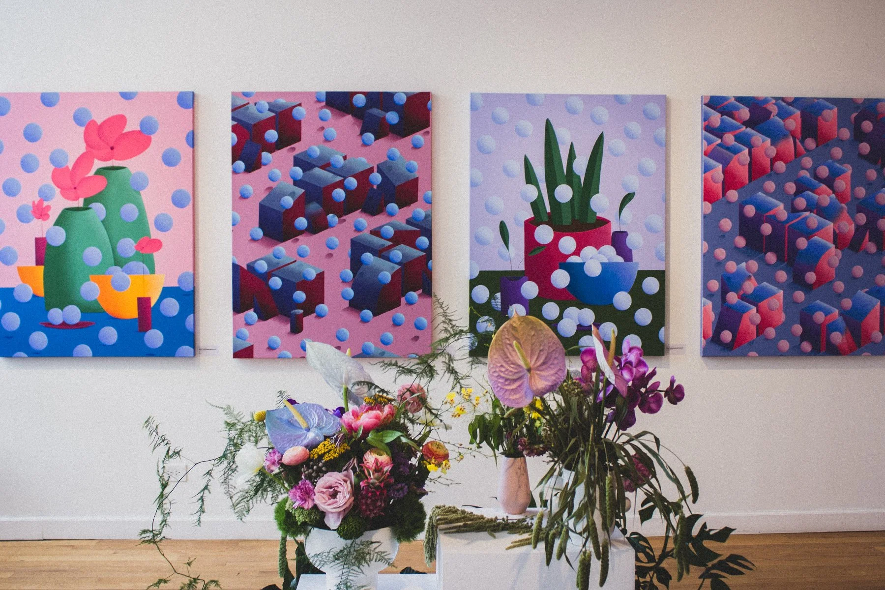 Gallery with four vibrant, abstract paintings featuring geometric and floral motifs, with a display of colorful flower arrangements in front.