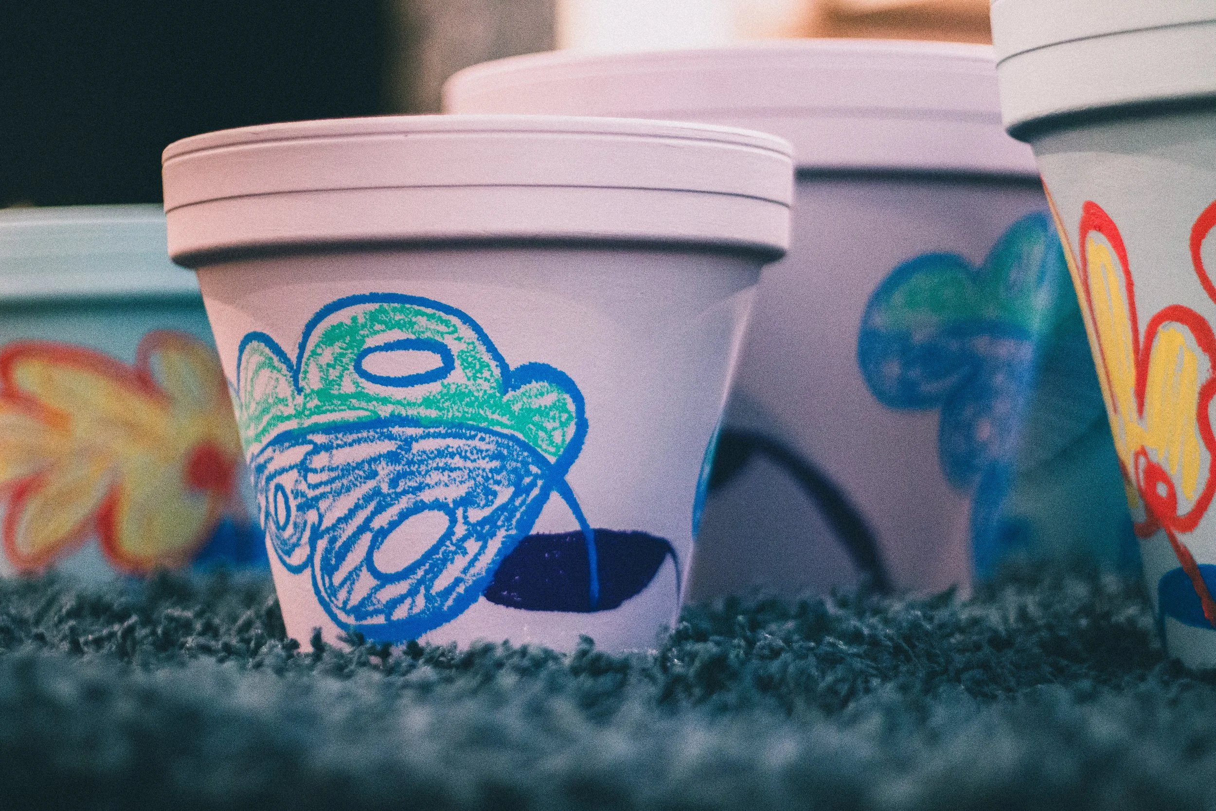 Colorful paper cups with childlike crayon drawings and designs, placed on a gray textured surface.
