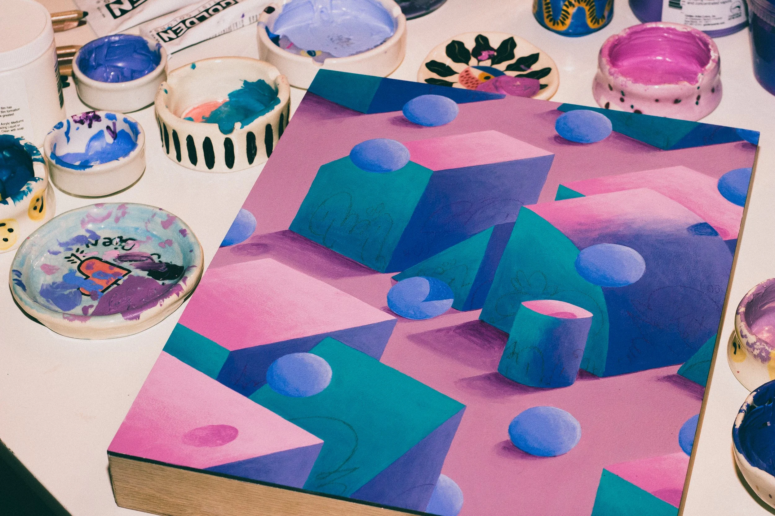 A colorful abstract painting featuring pink, purple, and blue geometric shapes, including cubes, cylinders, and spheres, surrounded by various open paint pots and brushes.