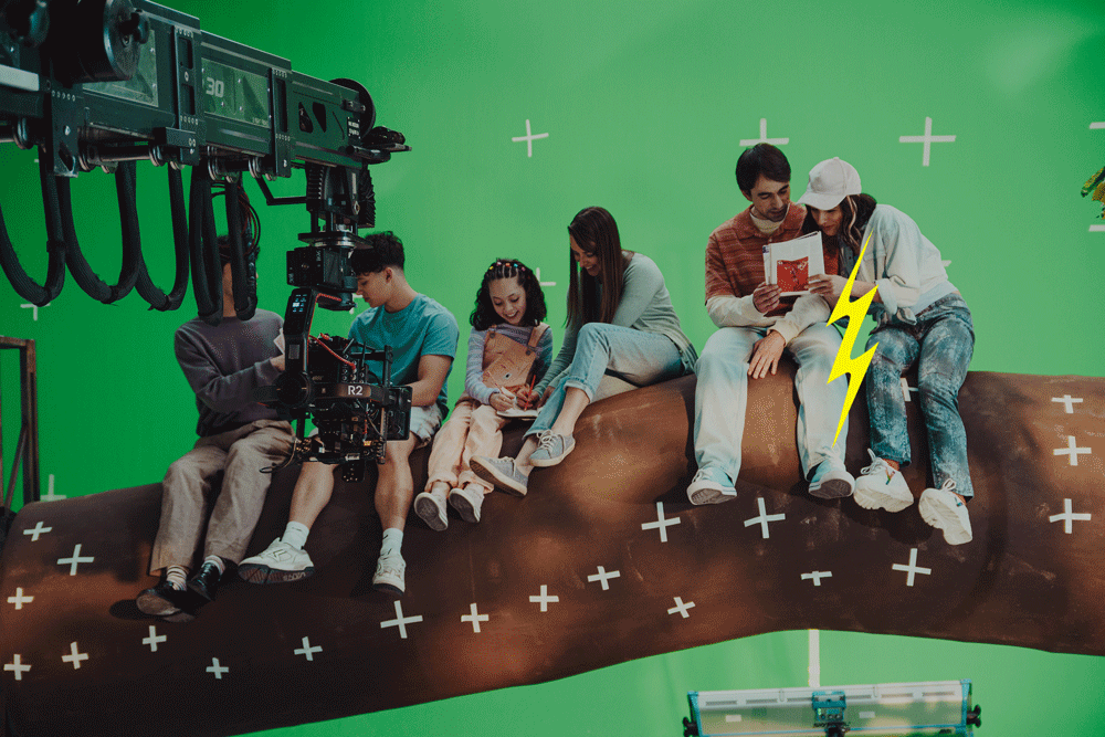 Children and young adults sit on a large fake tree trunk in front of a green screen while crew film on set.