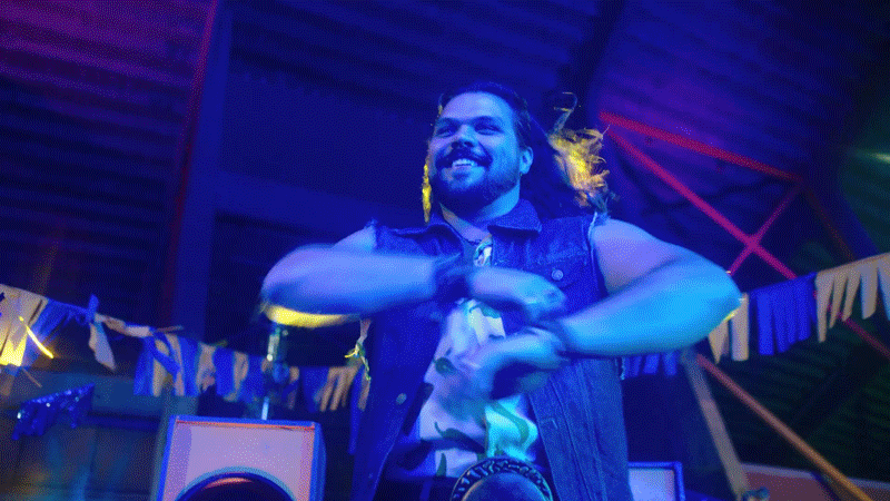 A man with long hair and a beard smiling, wearing a sleeveless denim vest, dancing with arms crossed in front of his chest in a colorful, lively setting with decorations and lights.