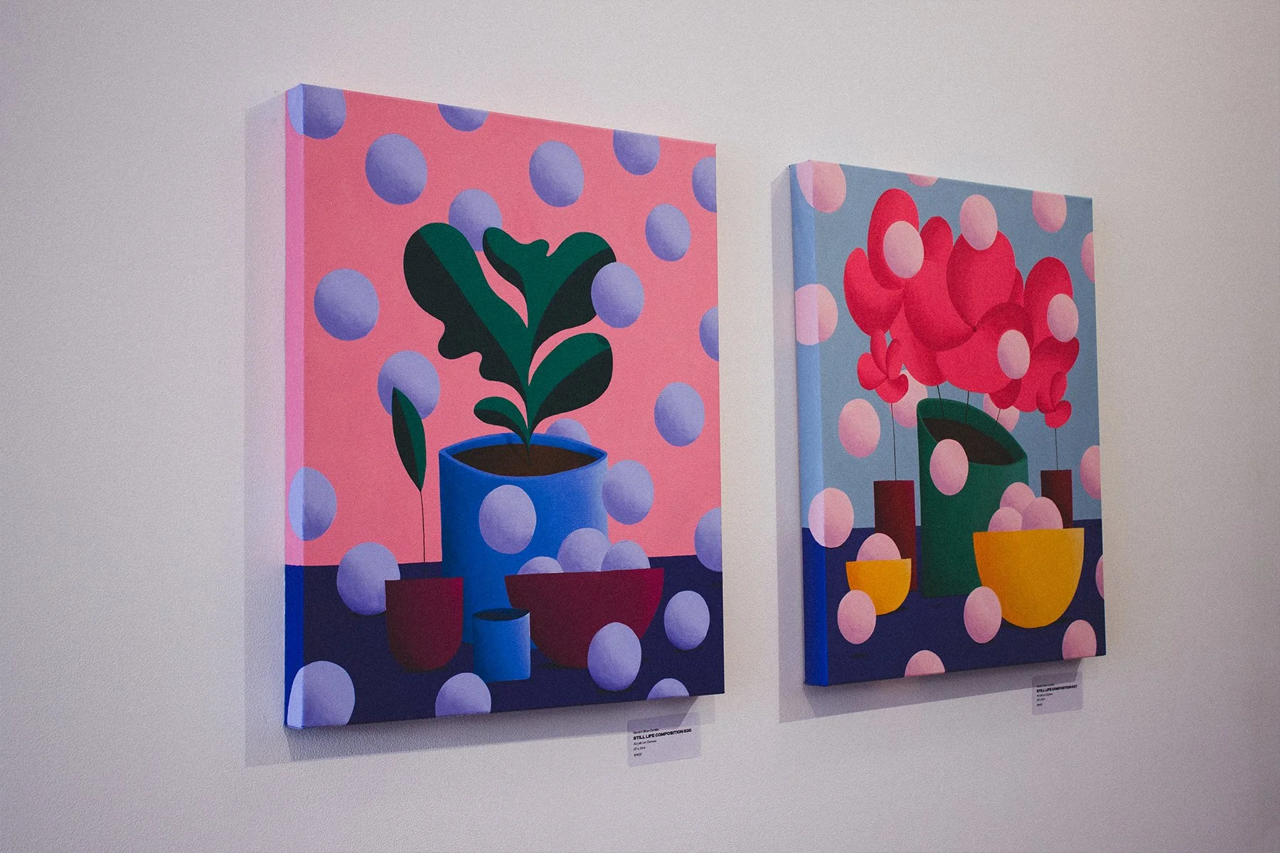 Two colorful paintings of potted plants with balloons in the background are displayed on a gallery wall.