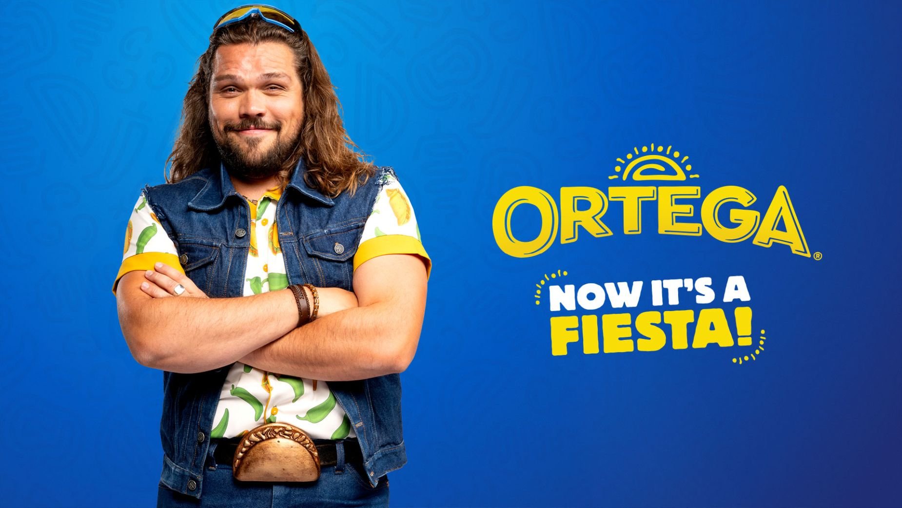 A man with long brown hair, a beard, and glasses on his head, wearing a denim vest over a colorful shirt with peppers, stands with his arms crossed and smiles. Behind him is blue background with the logo 'ORTEGA' and the text 'Now it's a Fiesta!'