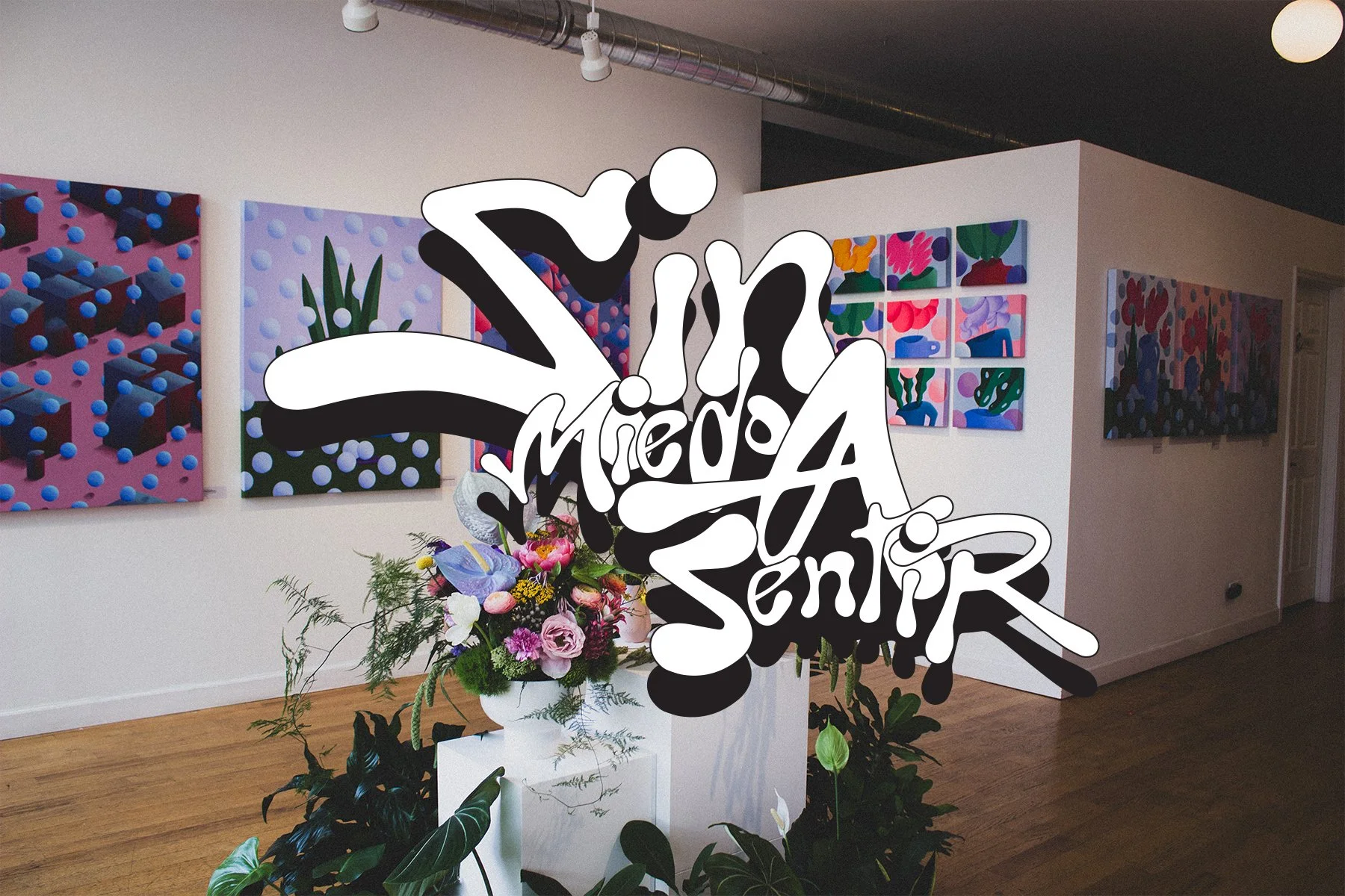 An art gallery with colorful paintings displayed on the walls and a floral arrangement in the center. Overlaid is stylized text that says 'Vive Medio A Sentir'.