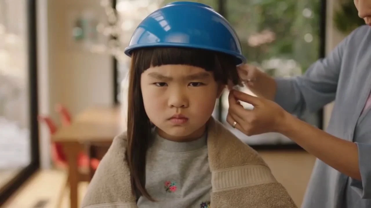 A young girl with straight dark hair wearing a blue safety helmet inside a room with large windows and a blurred background. A person, partially visible, is adjusting her helmet.