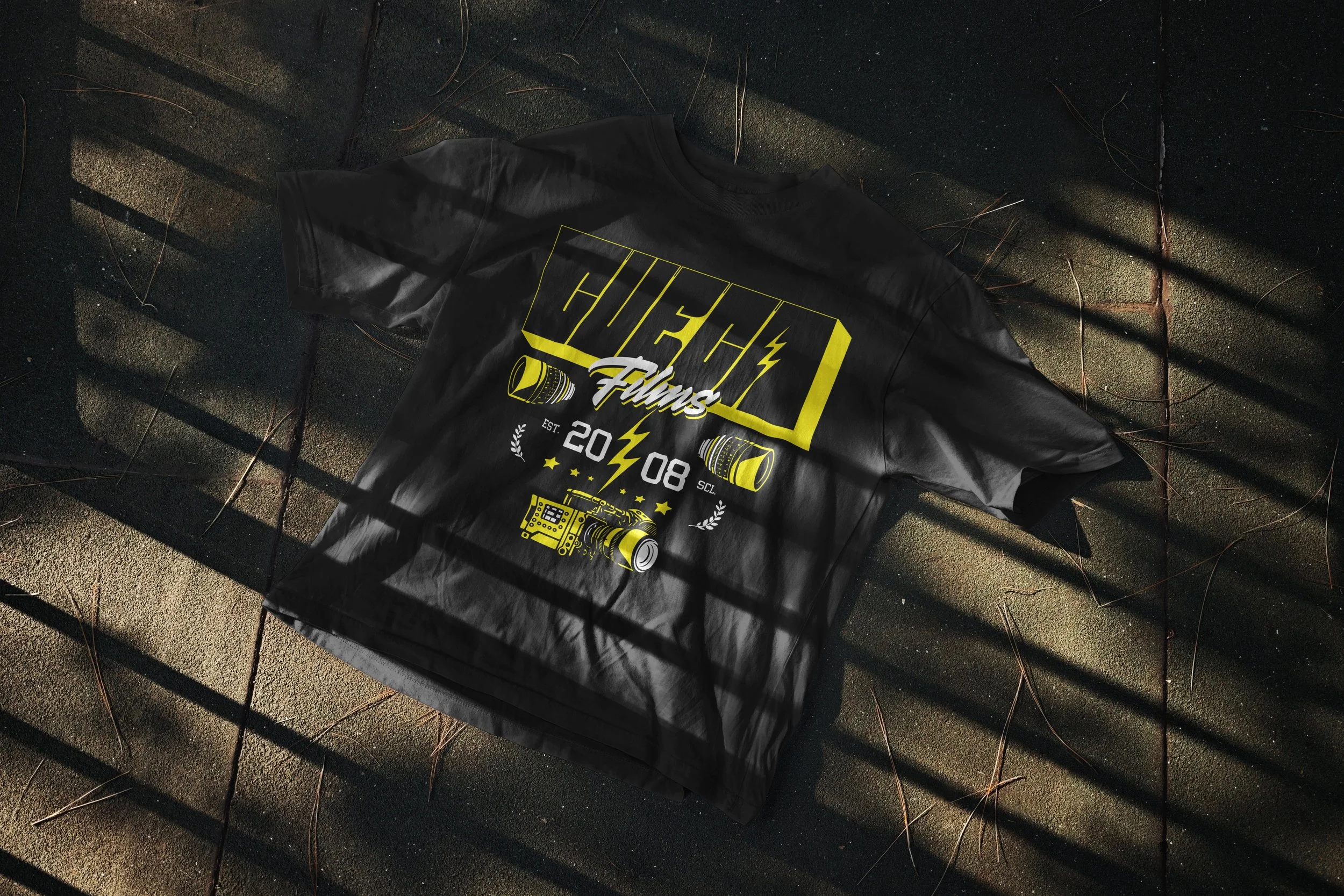 Black T-shirt with yellow graphic and text, lying on a textured pavement ground with shadows from nearby vertical bars.