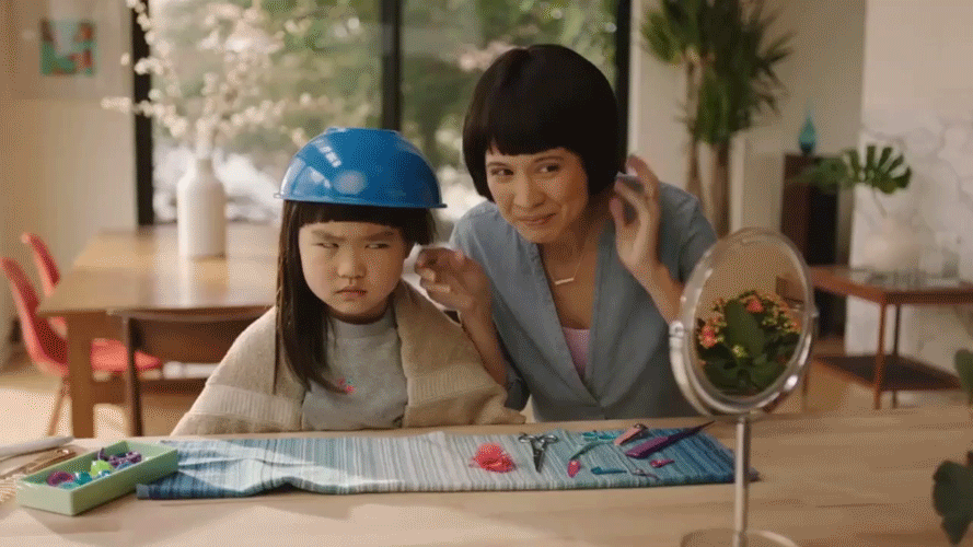 An adult woman helping a young girl with a craft project at a table, the girl wearing a blue helmet, in a bright room with plants and large windows.