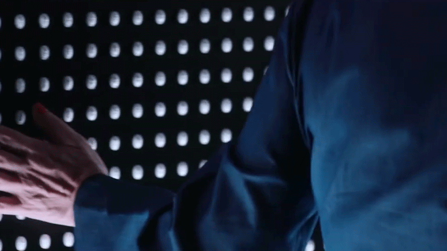 Close-up of a person wearing a dark blue shirt, pointing at a black wall with evenly spaced white circular dots.