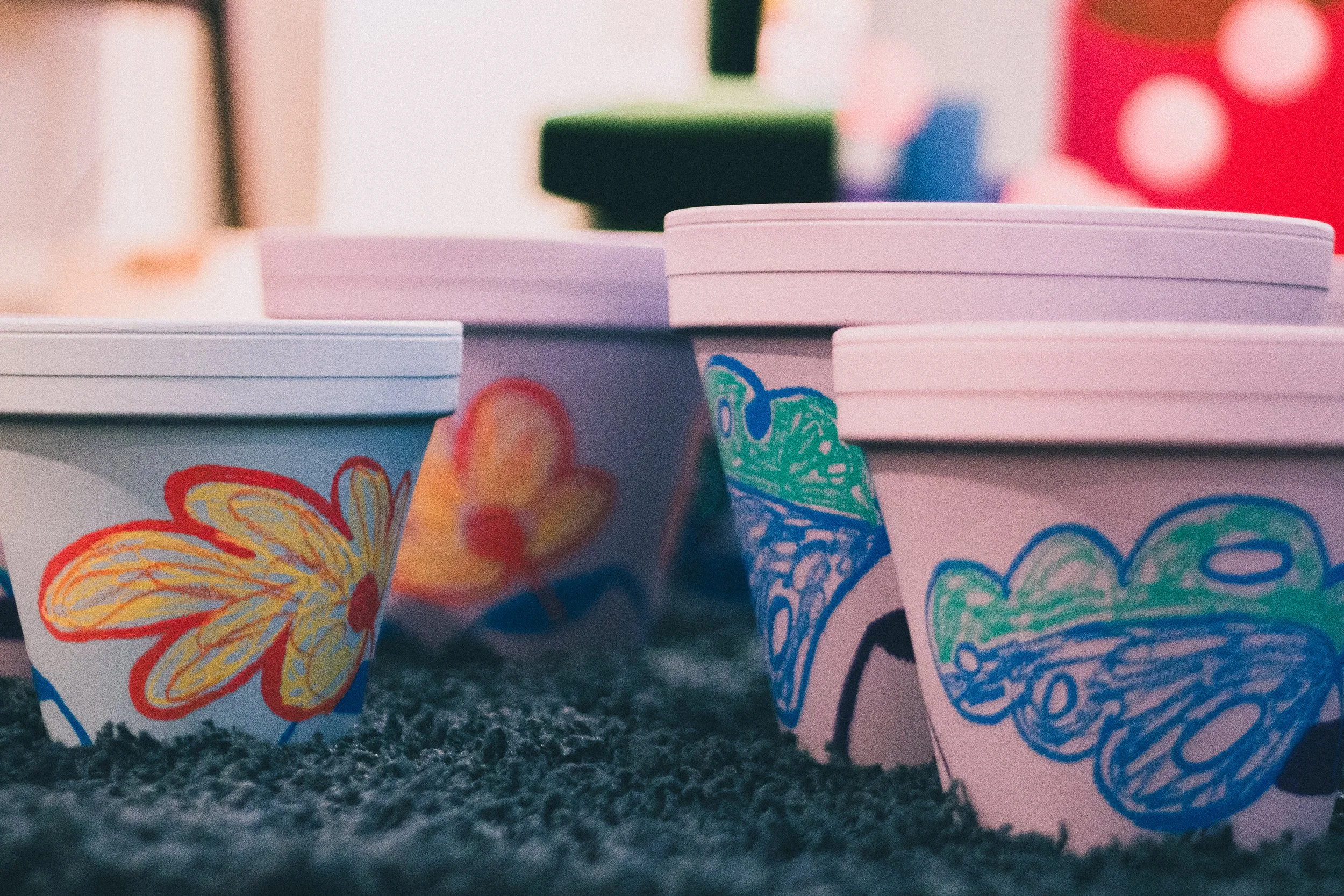 Colorful paper cups with children’s drawings, including a butterfly, a flower, a dinosaur, and a cloud, placed on a carpeted surface.