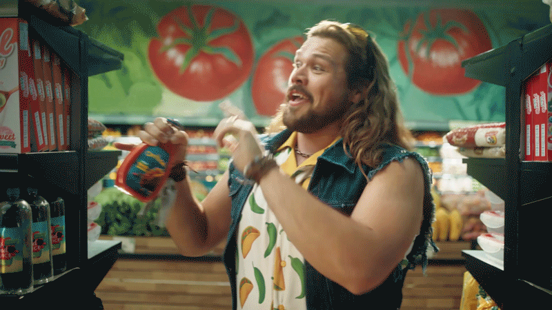 A man with long hair and a beard in a grocery store, wearing a denim vest and a fruit-patterned apron, holding a spray bottle while talking animatedly.