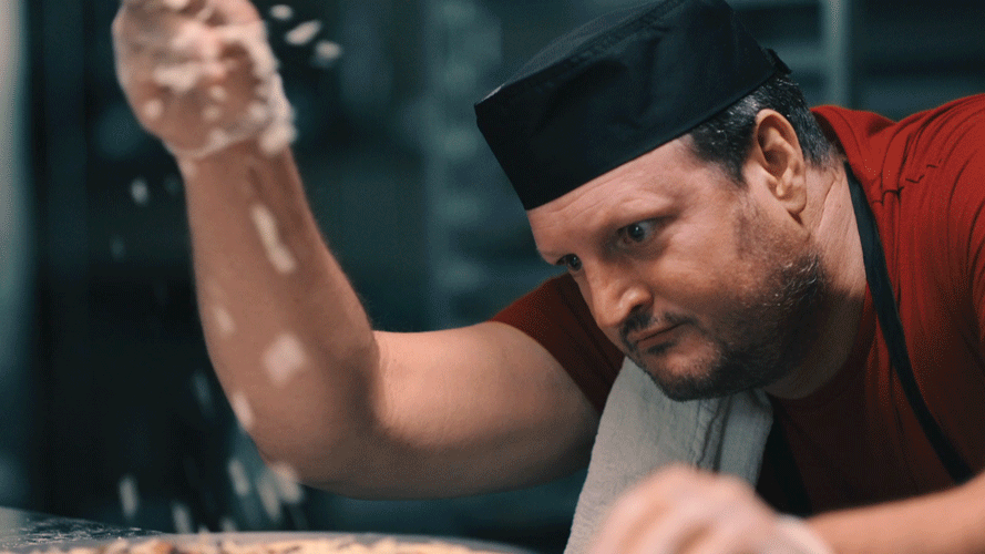 A chef wearing a black hat and red shirt is leaning over a surface, sprinkling flour.