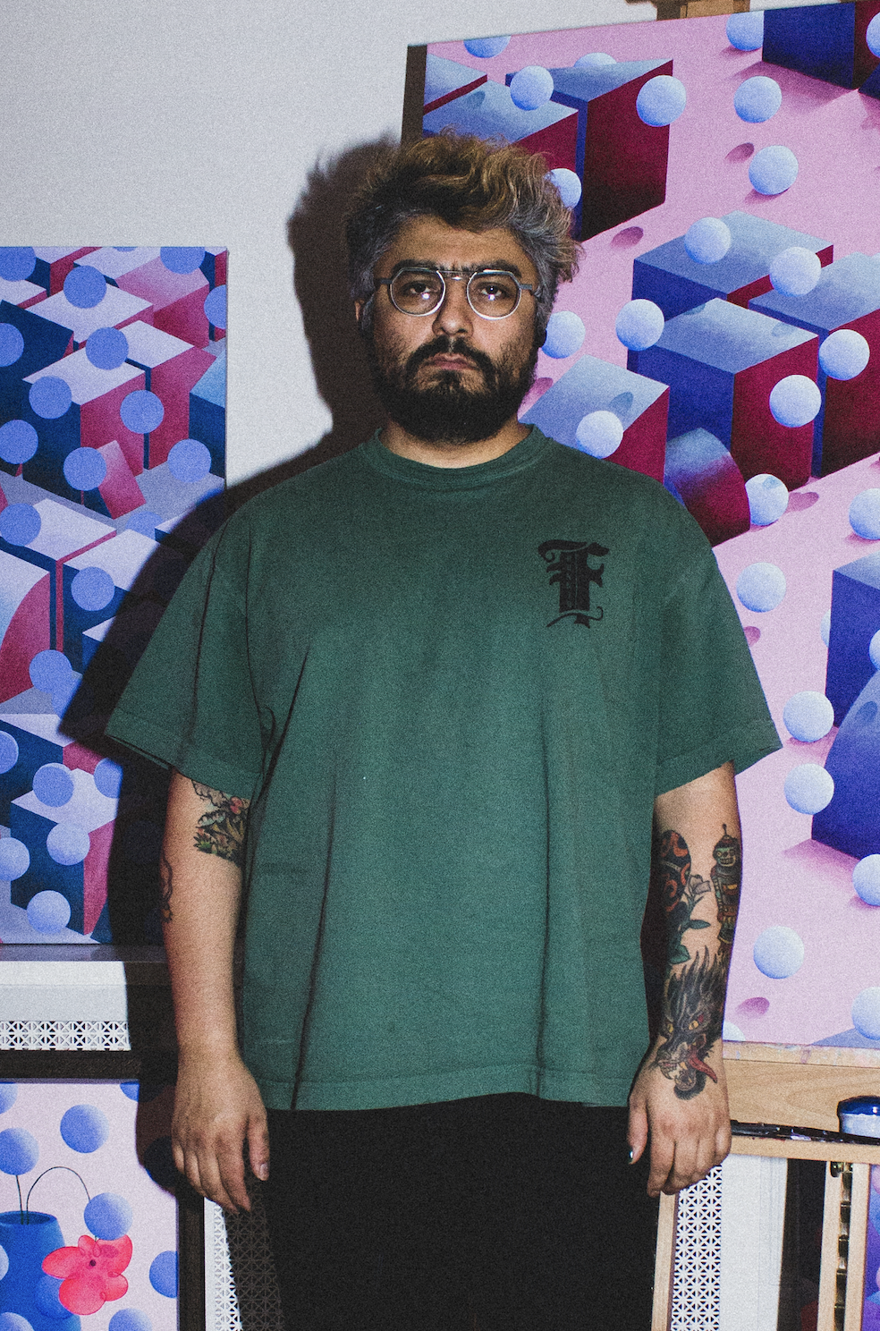 A man with glasses and tattoos standing in front of colorful abstract geometric paintings with cubes and spheres.