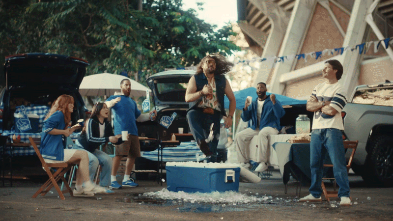 Group of friends at a tailgate party outdoors, with cars, coolers, and drinks, enjoying a lively gathering.
