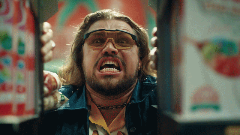 A man with long hair and a beard, wearing glasses and a dark jacket, looks distressed while standing between shelves of colorful snacks or cereal boxes.