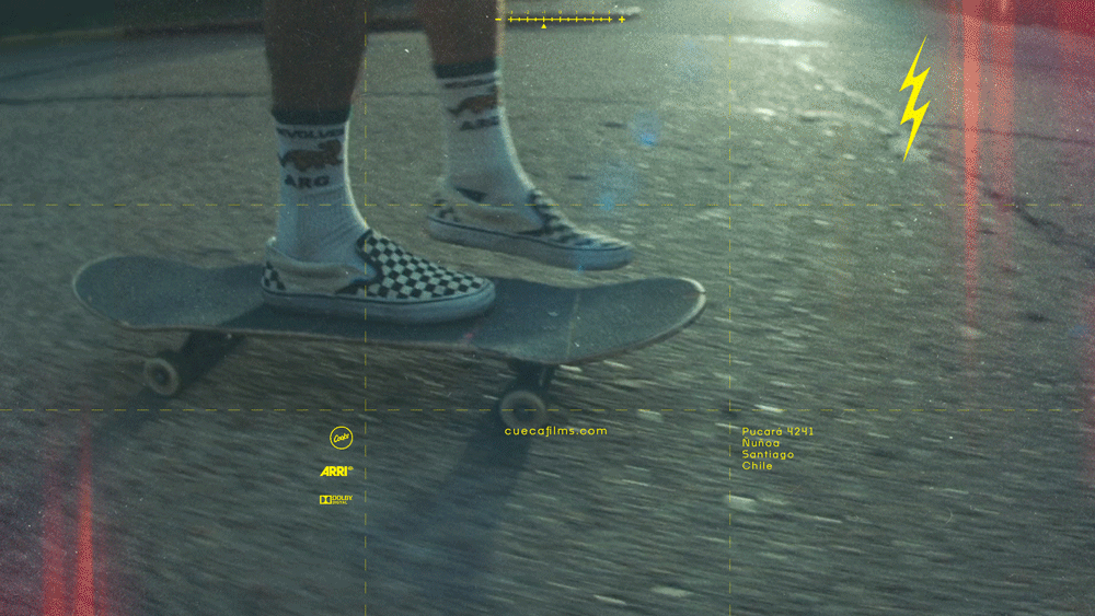 Close-up of a person skateboarding on a paved street, wearing checkered slip-on shoes, patterned socks, and casual clothing. The photo has a yellow overlay with text and symbols, including a lightning bolt and various logos.
