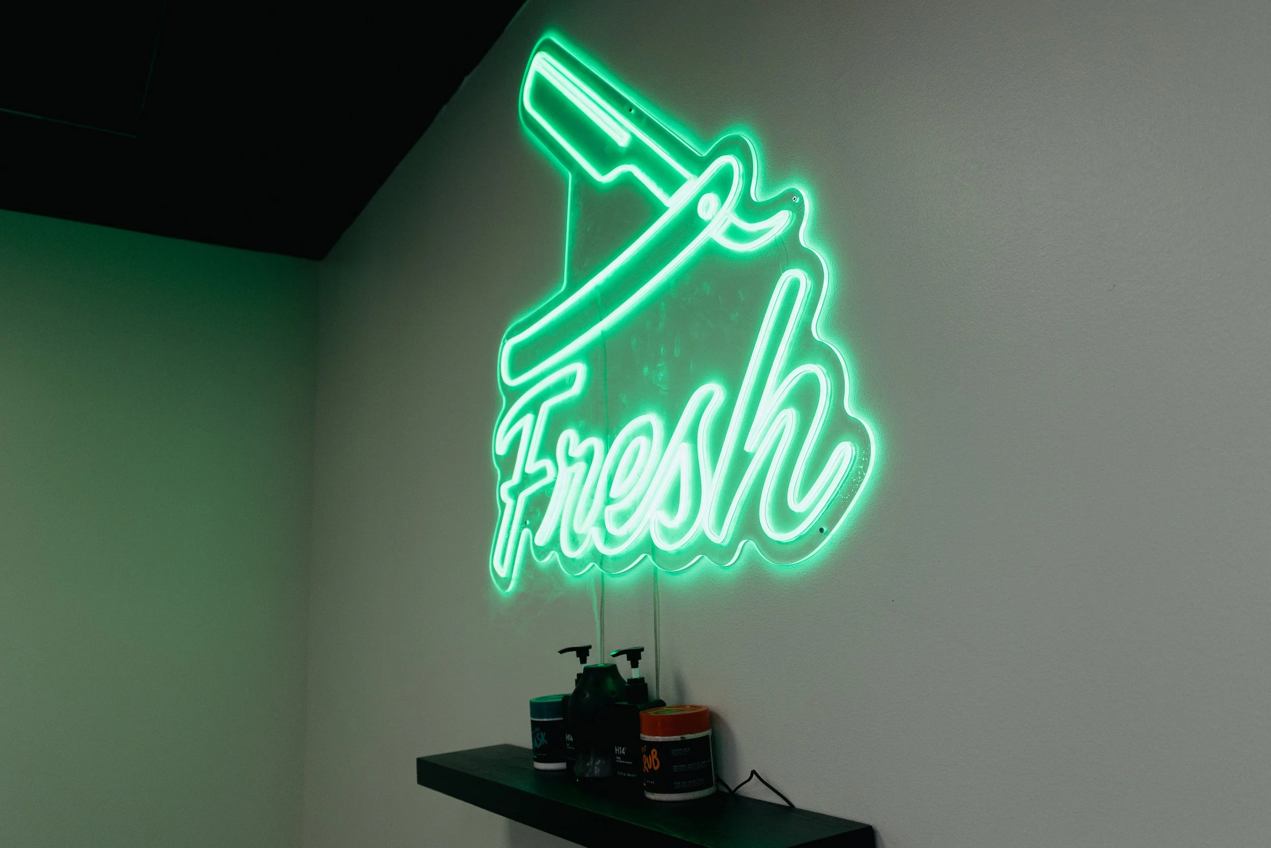 Green neon sign with a syringe and the word 'Fresh' on a wall above a black shelf with bottles and containers.