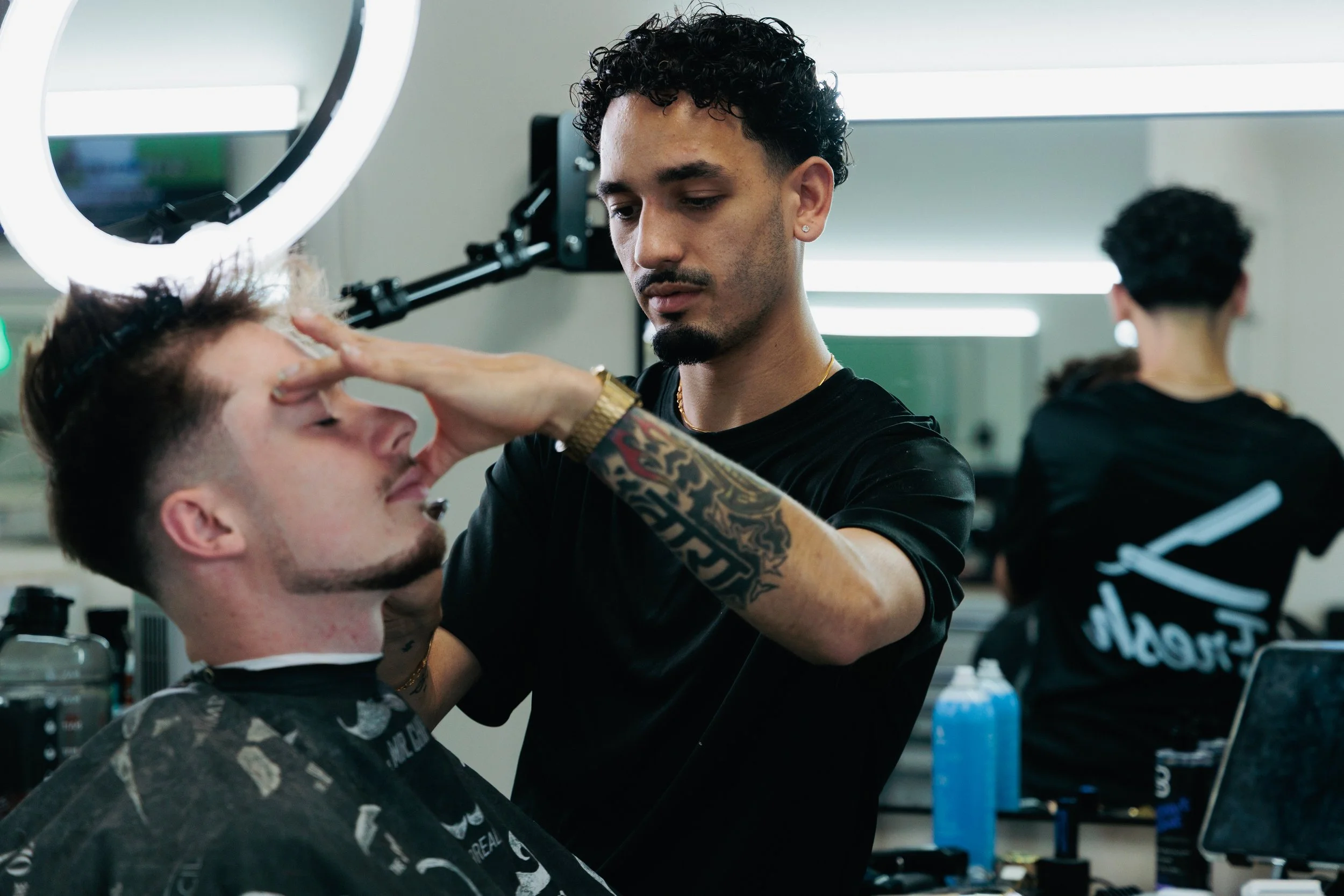Barber applying shaving cream to a man's face in a modern barbershop with a round mirror and bright lighting.