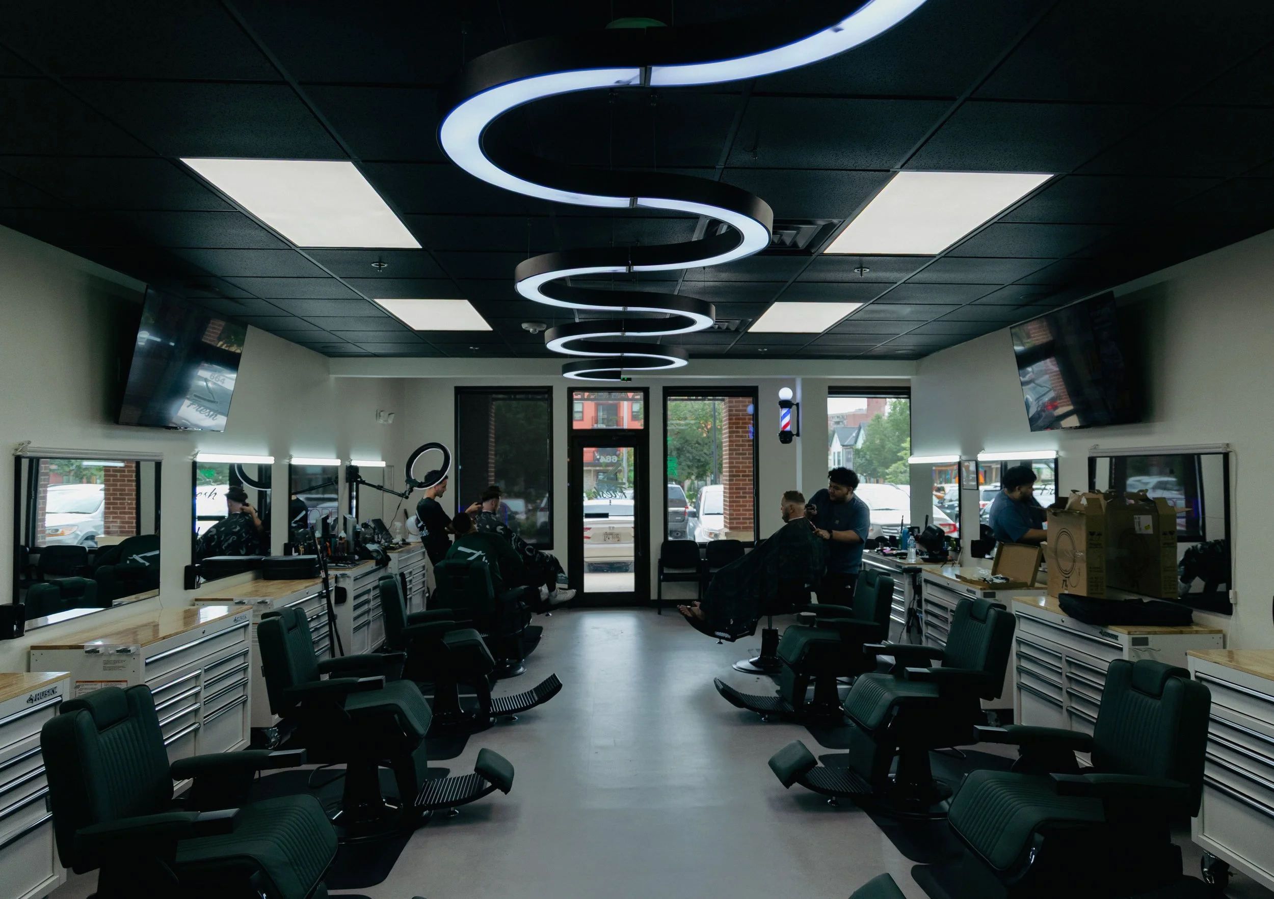 Barbershop interior with chairs, mirrors, and customers getting haircuts, illuminated by modern spiral ceiling light.