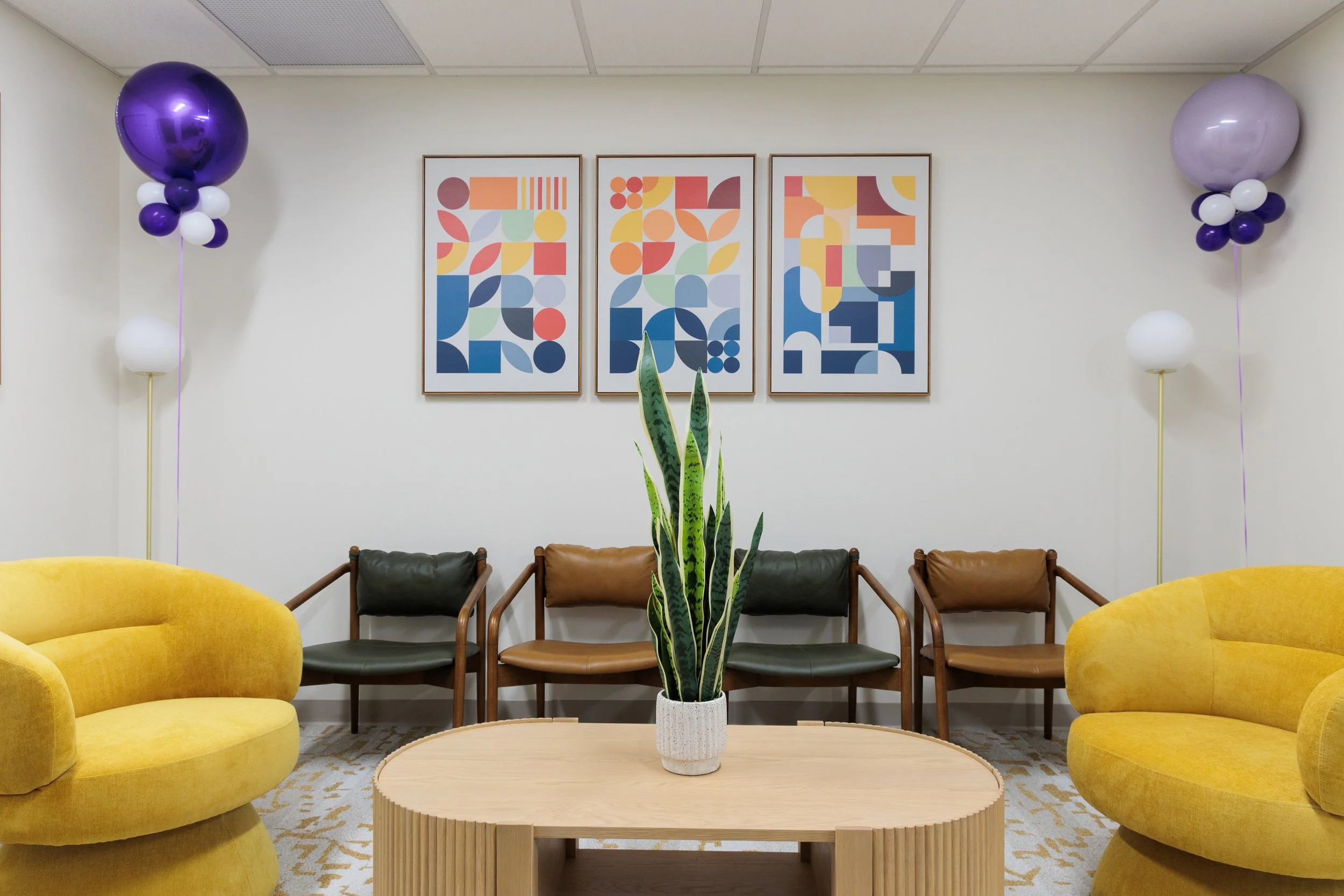 A brightly colored waiting room with yellow couches, a round wooden coffee table with a potted snake plant, abstract geometric artwork on the wall, and purple and white balloons on either side.