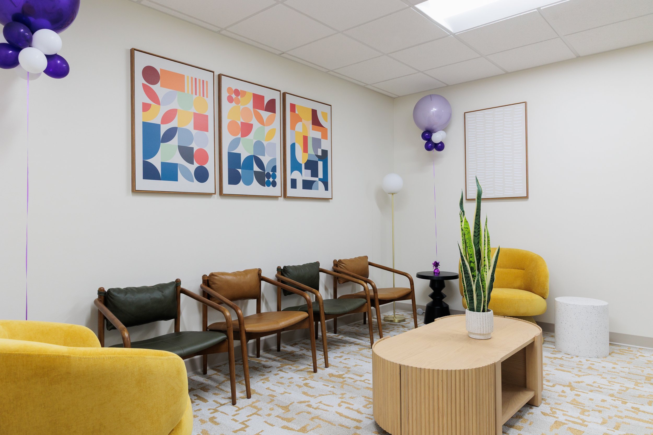 Waiting room with four chairs, a yellow sofa, a coffee table with a potted plant, balloons, and framed geometric artwork on the wall.