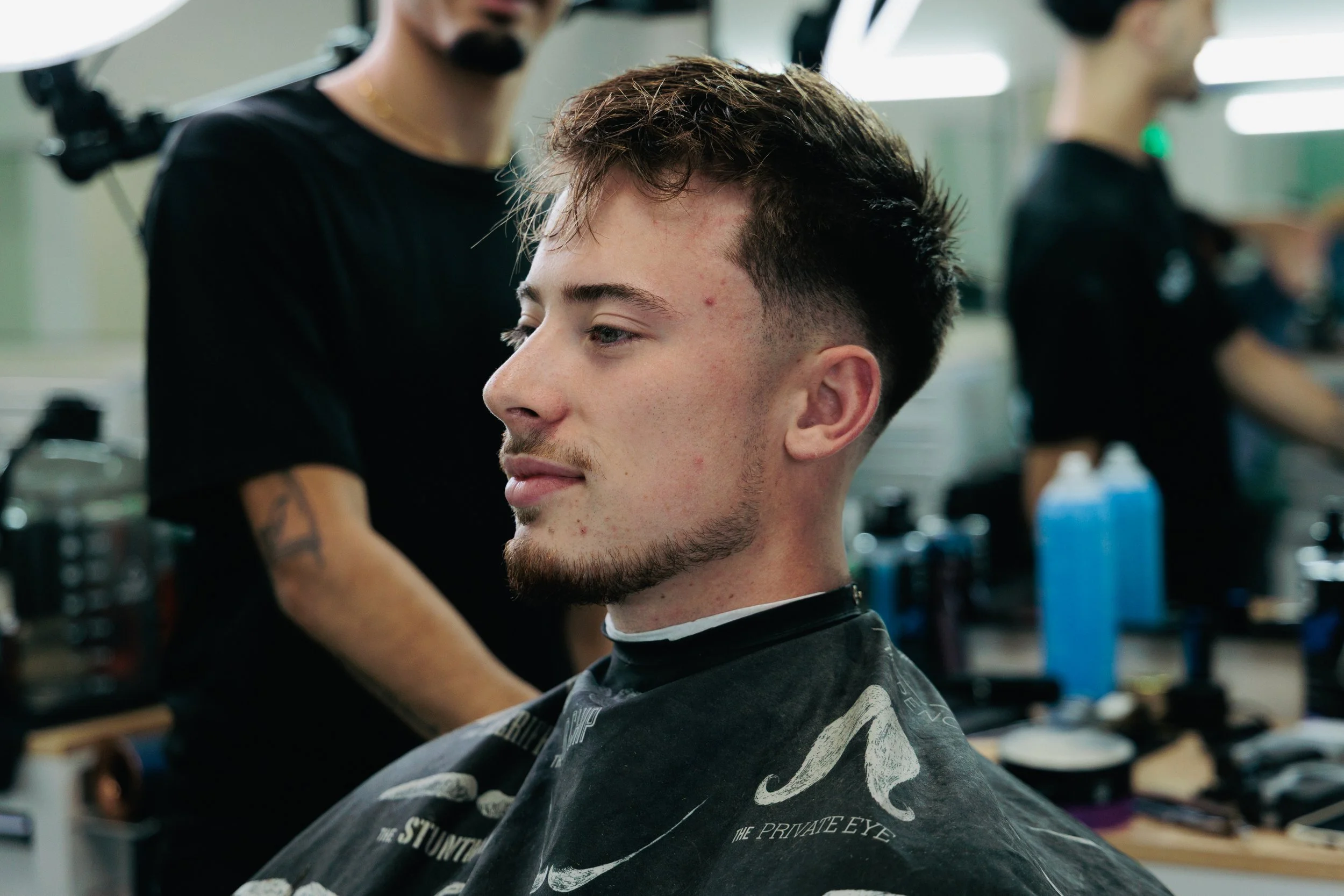 Young man with a fade haircut and beard sitting in a barber's chair, receiving a haircut. Barber is standing behind him, working on his hair, in a busy barbershop with various hair products and tools visible.