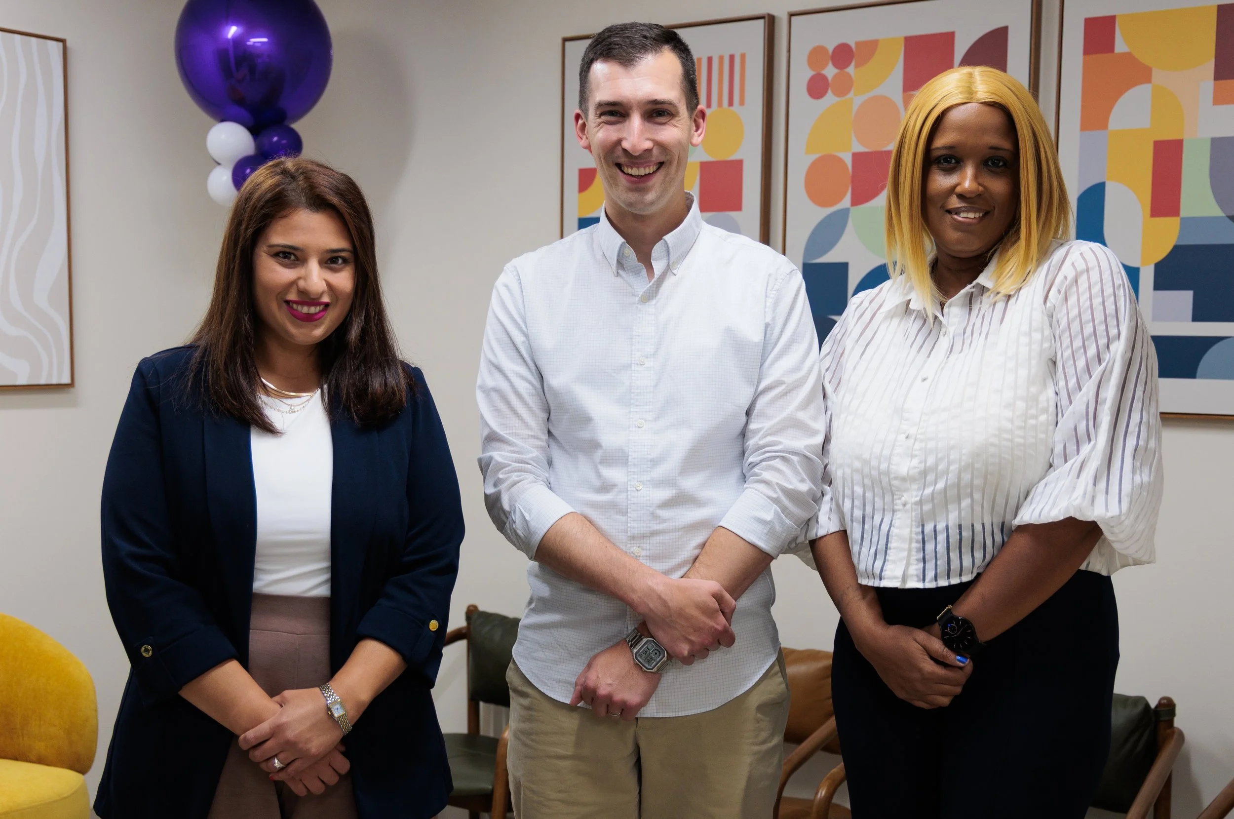 Three diverse professionals standing together and smiling in an office with colorful modern artwork on the wall.