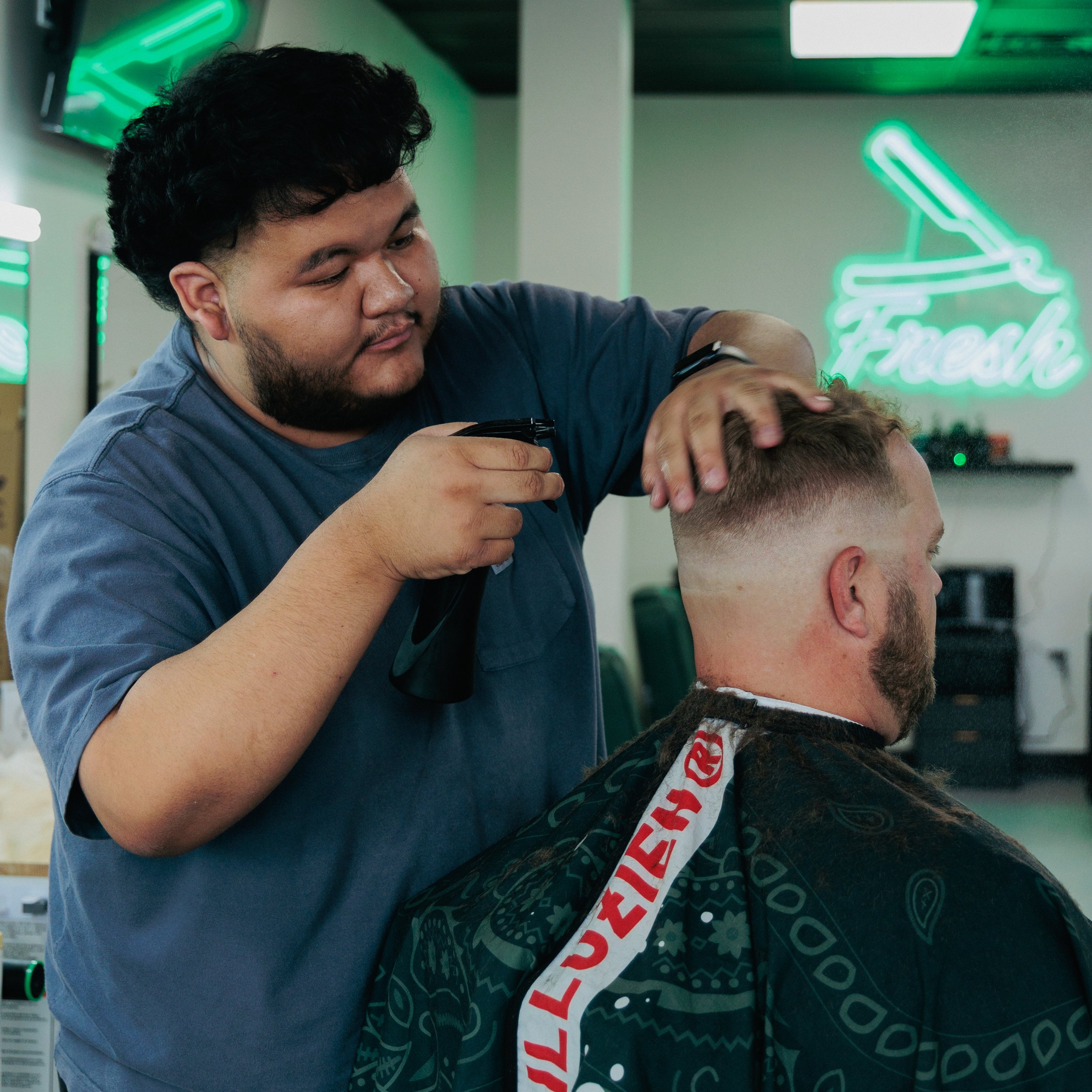 A man giving a haircut to another man in a barbershop, with neon signs and hair styling equipment visible in the background.