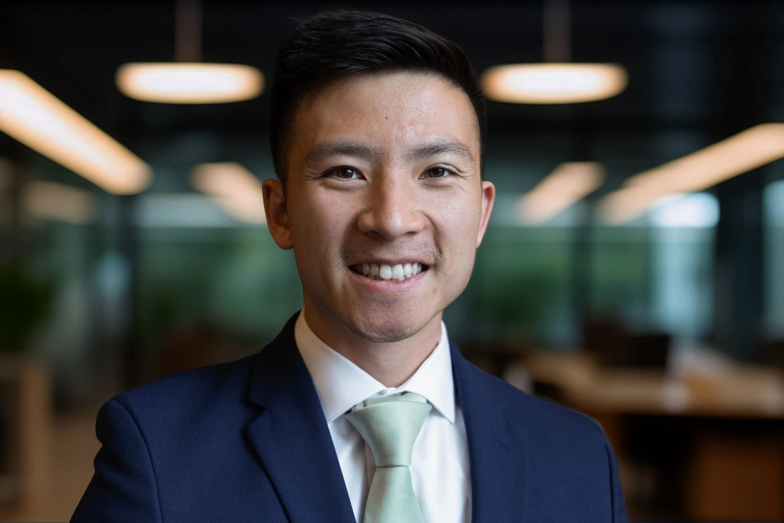 Headshot of a young man in a navy suit and light green tie, smiling with a blurred modern office background.