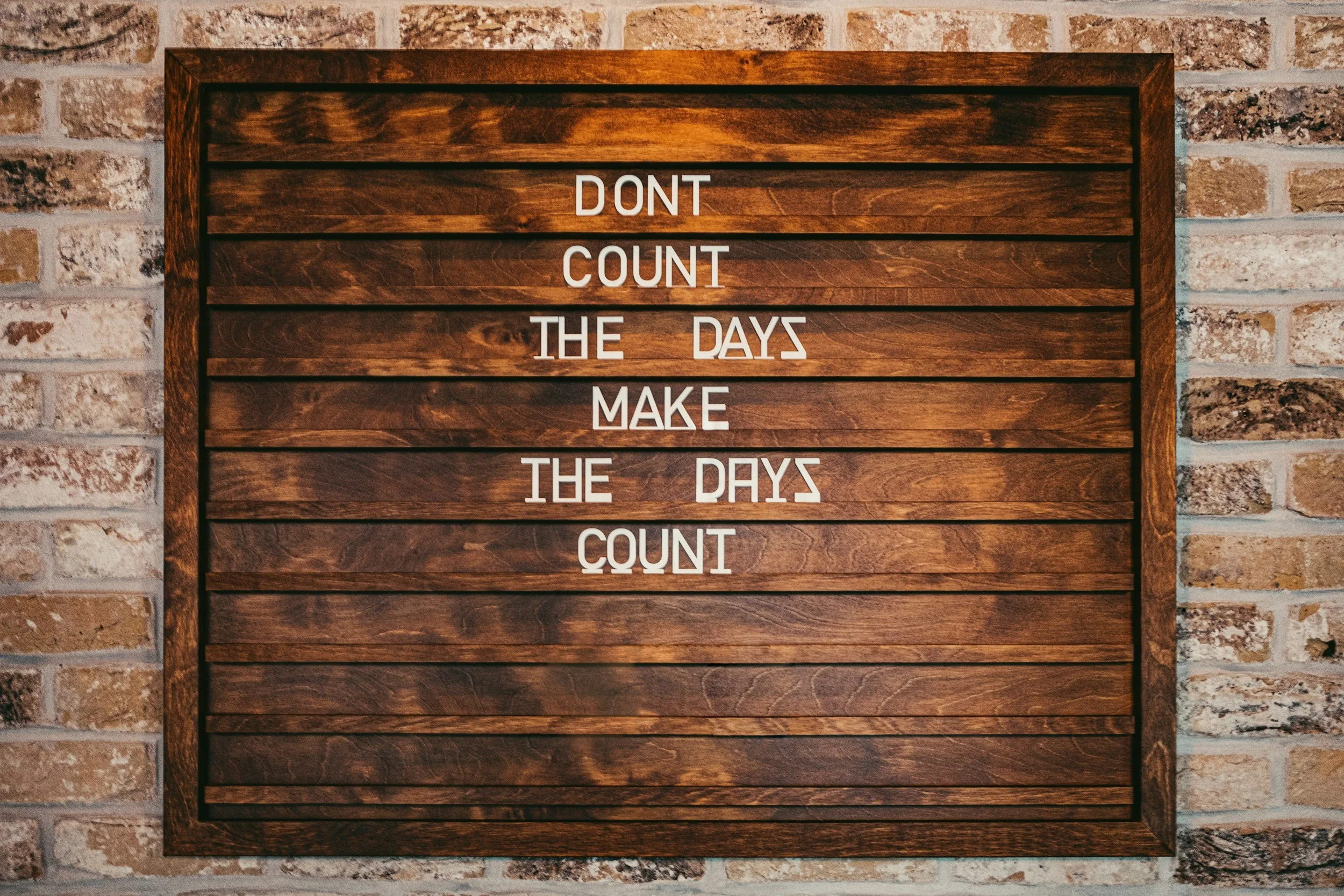 Wooden framed board with an inspirational quote: "Don't count the days, make the days count" against a brick wall background.
