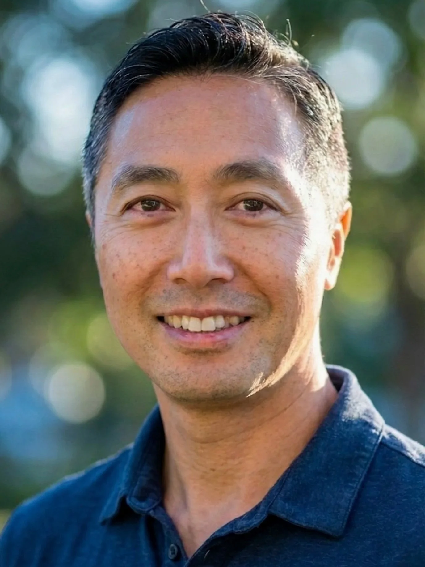 Close-up of a smiling man with short black hair, wearing a navy blue shirt, outdoors with a blurred background of trees and sunlight.